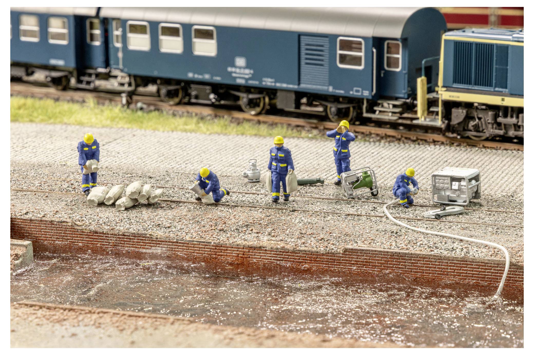 Szene zur Instandhaltung einer Miniatureisenbahn mit winzigen Arbeitern in gelben Helmen und blauen Uniformen, die das Gleis neben einem blauen Personenzug reparieren.