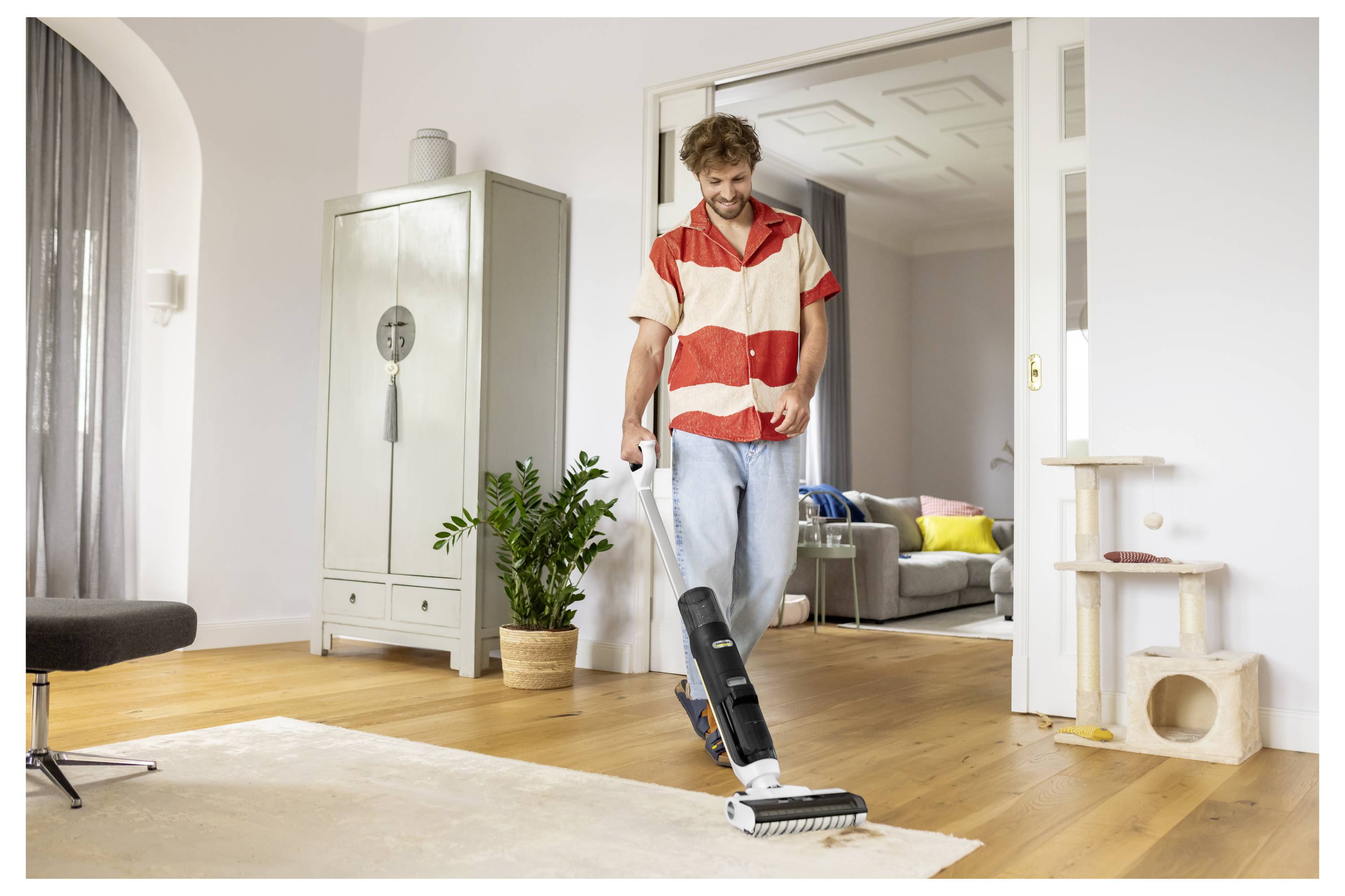 Ein Mann mit einem Staubsauger in einem Wohnzimmer mit Holzböden, der einen Staubsauger auf einen Teppich zubewegt, in einem hell erleuchteten Wohnrauminterieur.
