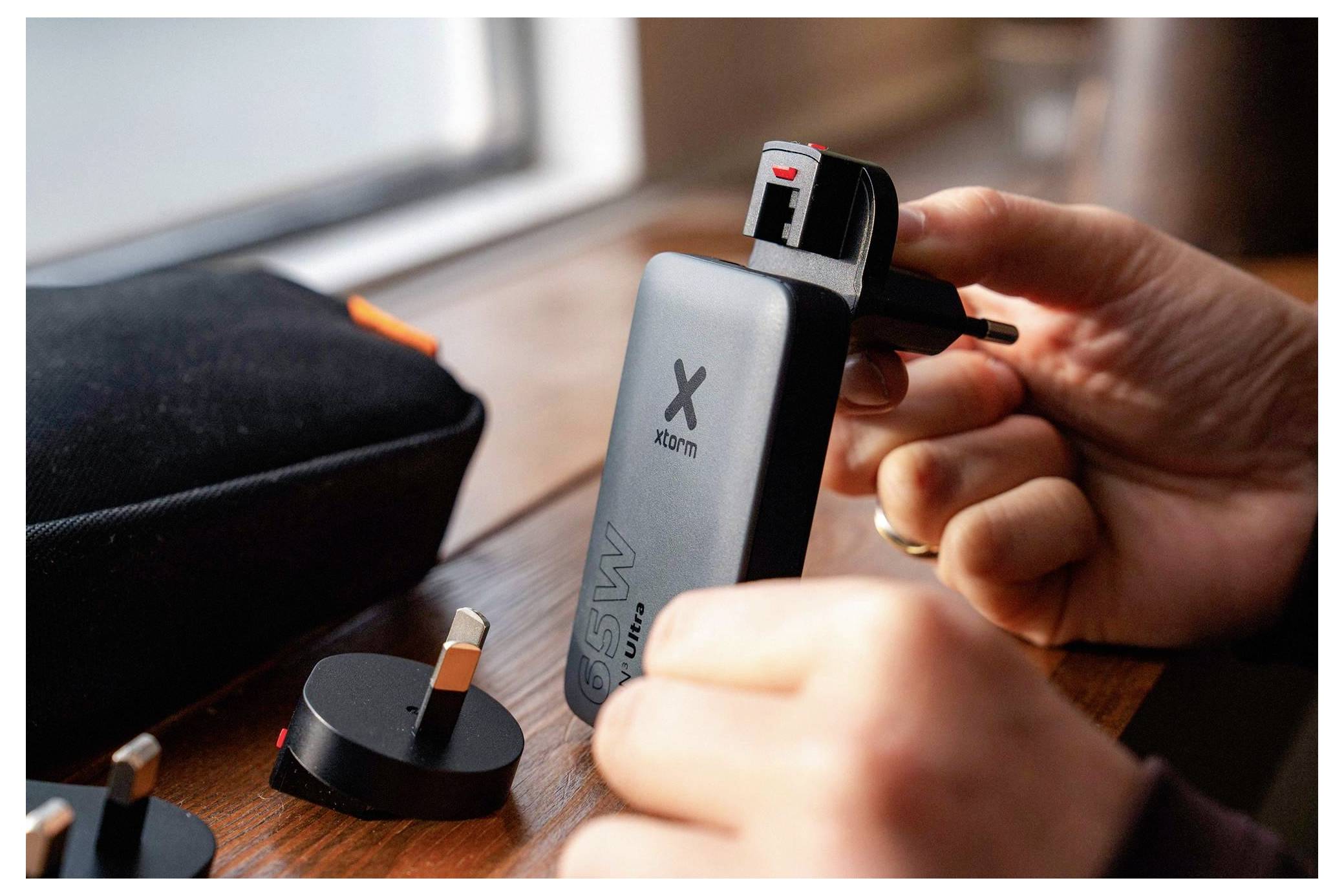 Hände, die einen britischen Adapter in eine tragbare Xiaomi-Powerbank auf einem Tisch einstecken, mit einem Ladeetui und anderen Steckern in der Nähe.