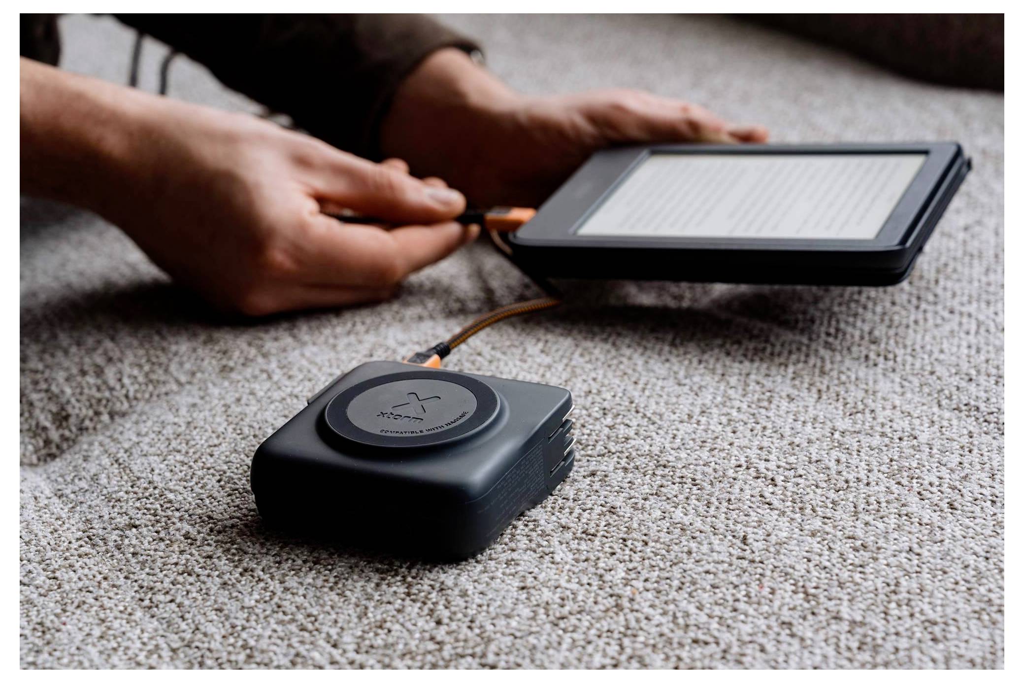 Eine Person lädt einen E-Reader mit einer tragbaren Powerbank über ein Kabel auf einem Teppichboden auf.
