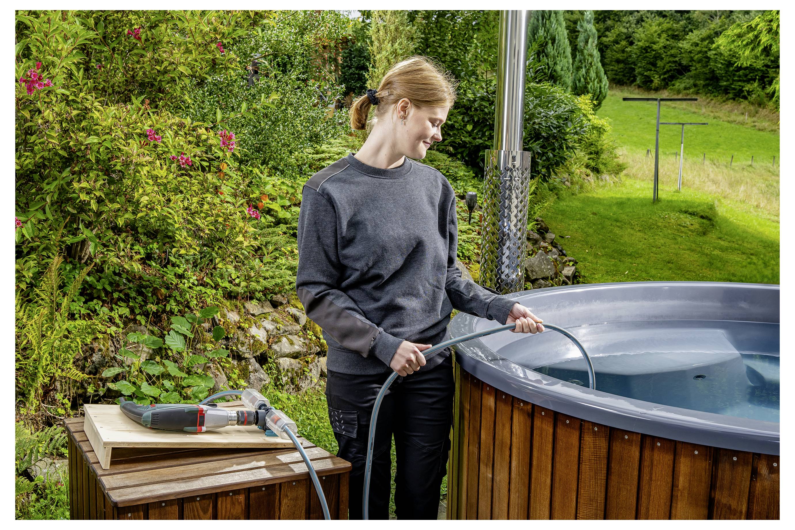 Eine Frau füllt eine hölzerne Badewanne im Freien mit einem Gartenschlauch neben einem Tisch mit Werkzeugen in einem Garten.