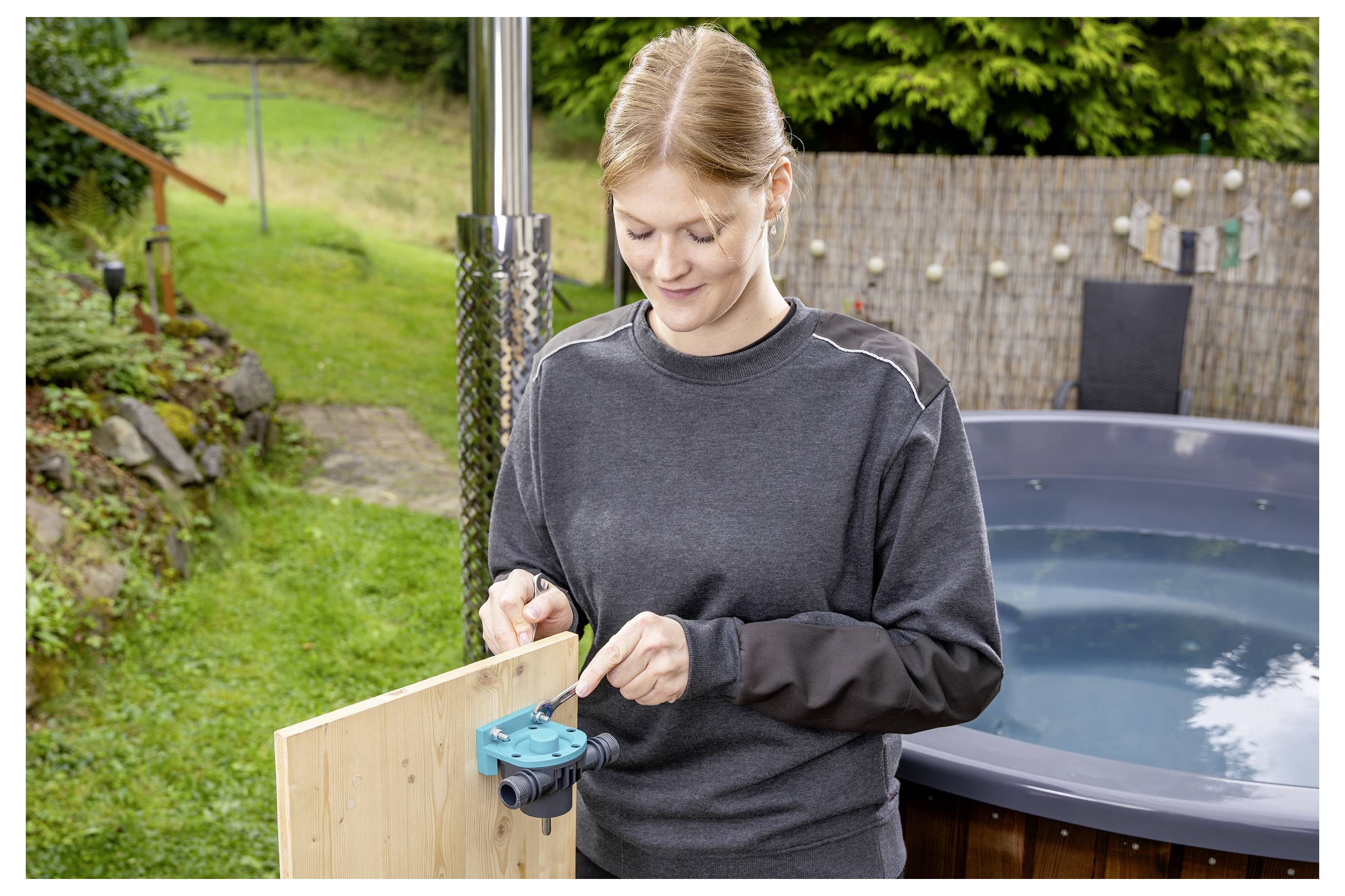 Frau, die draußen neben einem Whirlpool ein Holzpaneel montiert, dabei ein blaues Spannwerkzeug verwendend.