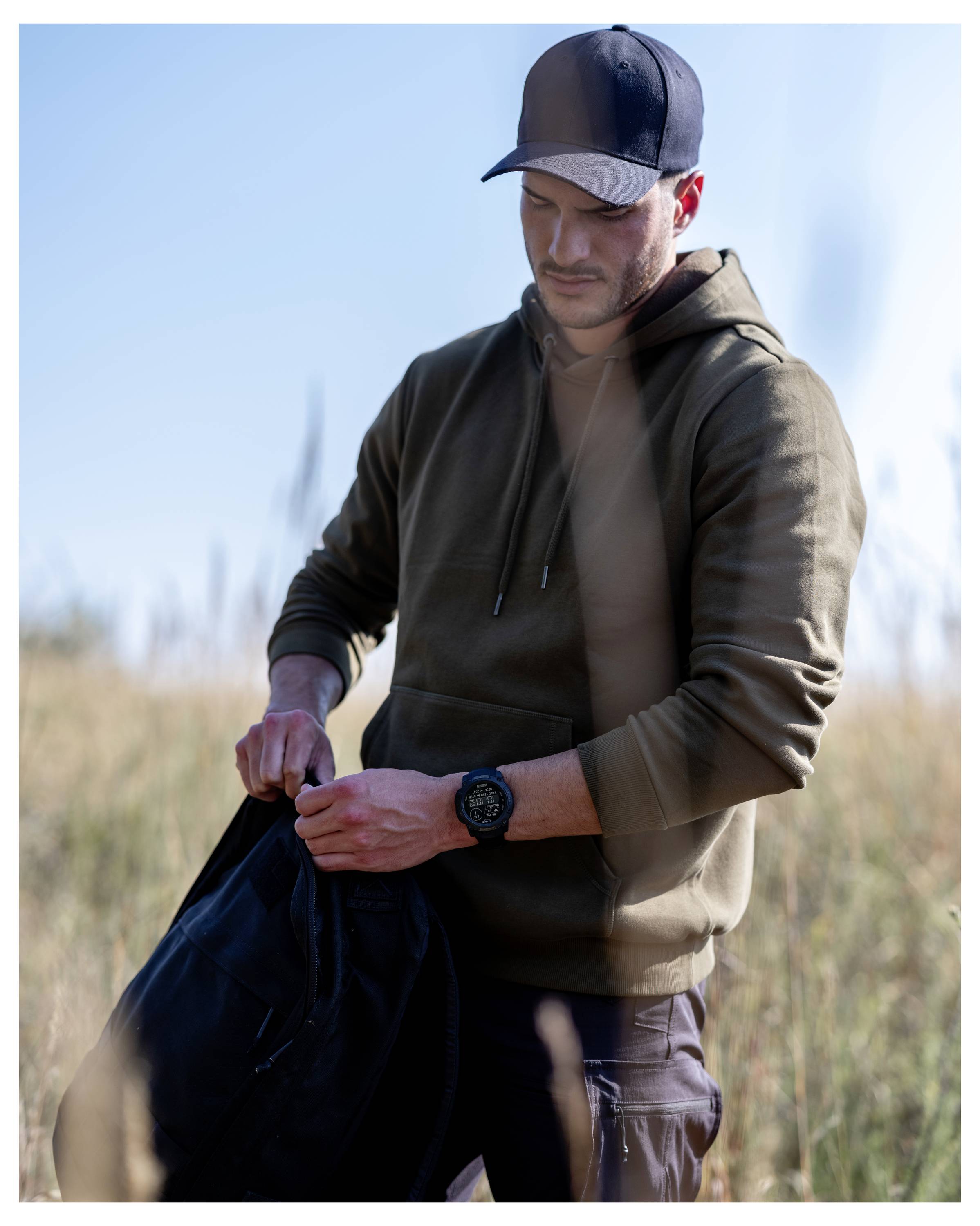 Mann im Freien auf einem Feld, trägt eine Mütze und einen Hoodie, schaut nach unten während er eine schwarze Tasche oder Jacke hält.