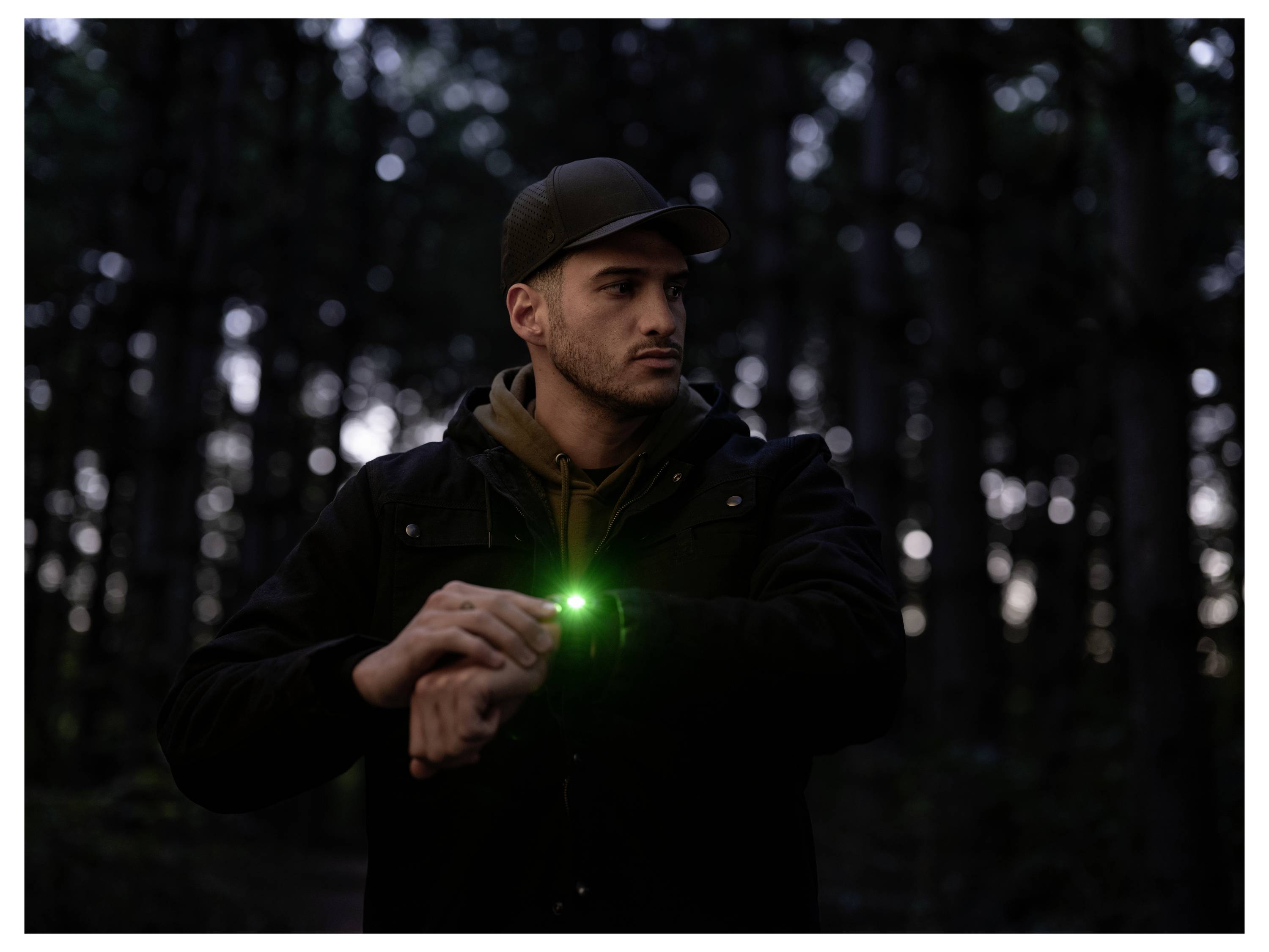 Mann in einem dunklen Wald, der bei Einbruch der Dunkelheit auf seine Armbanduhr schaut, mit einem grünen Licht, das von seiner Uhr oder seinem Gerät leuchtet.