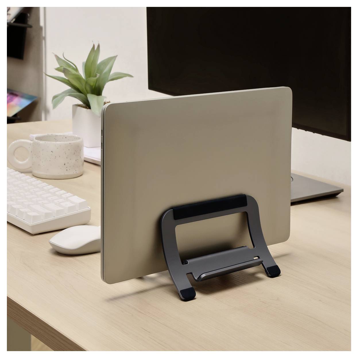 Ein geschlossener silberner Laptop steht aufrecht auf einem schwarzen Ständer auf einem Holzschreibtisch neben einer Tastatur, einer Maus, einer Tasse und einem Monitor.