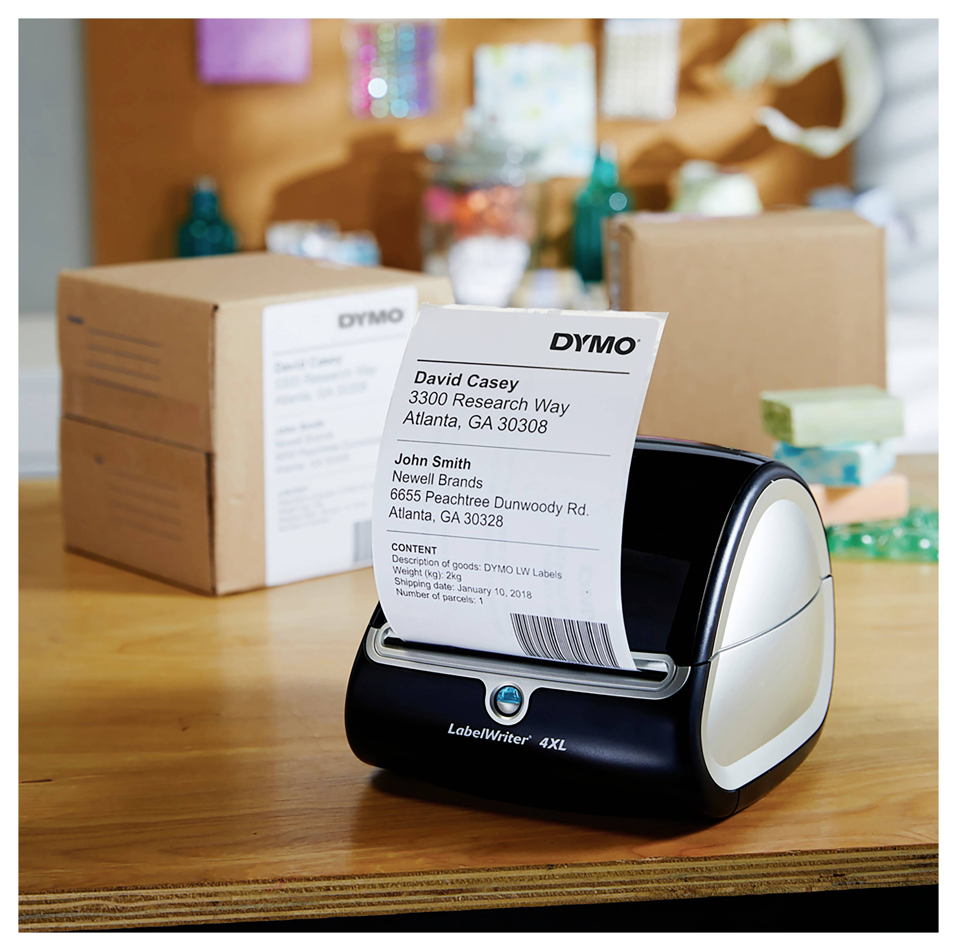 DYMO LabelWriter 4XL druckt ein Versandetikett mit Empfängeradressen auf einem Schreibtisch, mit gepackten Kartons im Hintergrund.