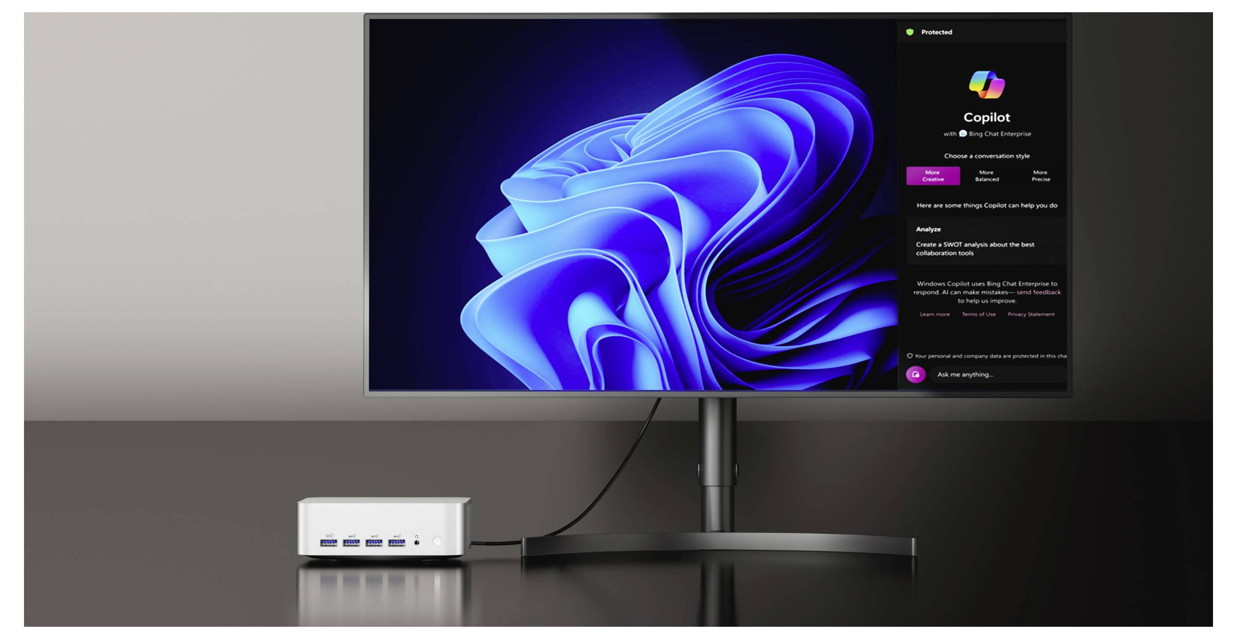 Ein Desktop-Monitor zeigt die Windows 11 Copilot-Oberfläche neben dem blauen Windows-Hintergrundbild an, mit einem kleinen Docking-Hub auf dem Schreibtisch darunter.
