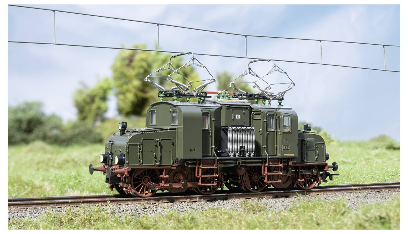 Eine grüne Modell-Elektrolok auf Gleisen, mit angehobenen Stromabnehmern unter Oberleitung in einer grünen Außenanlage.