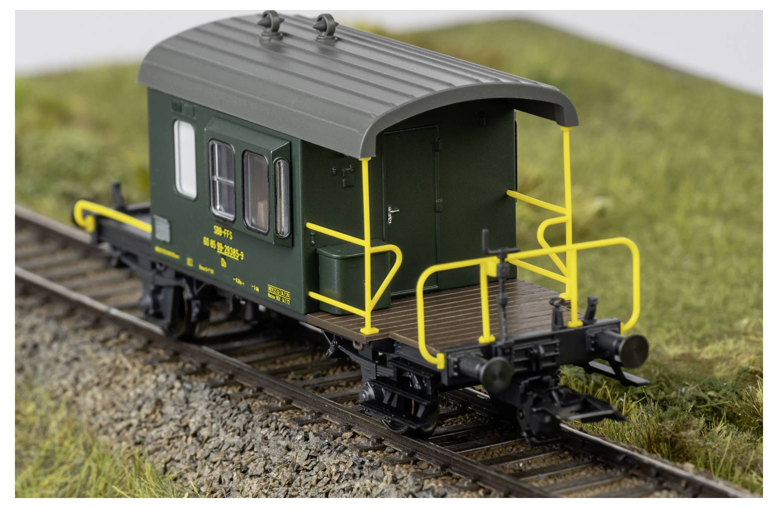 Grüner Modell-Güterwagen mit gelben Geländern auf Gleisen in einer Gras-Miniaturlandschaft.