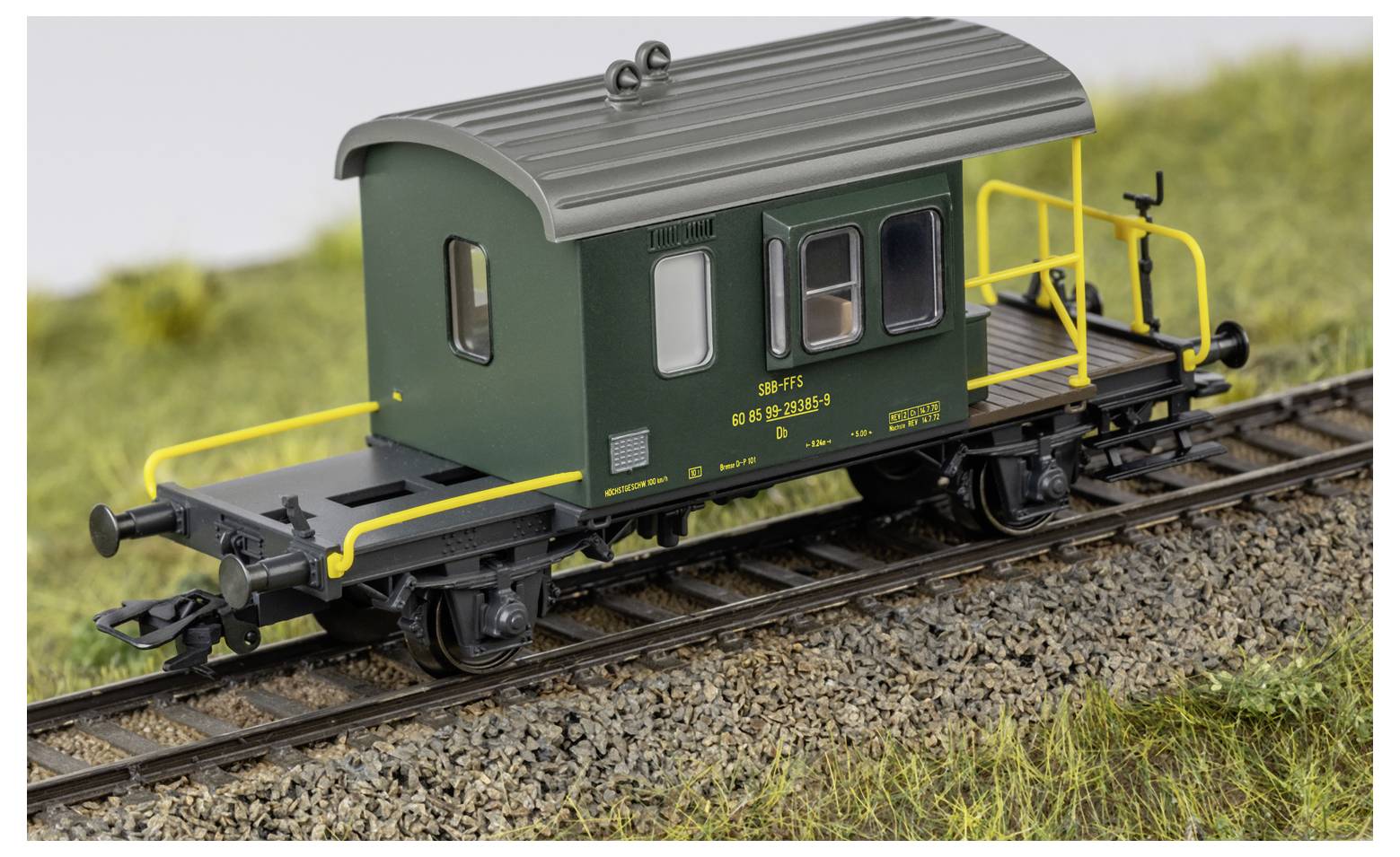 Ein Modell-Eisenbahn-Güterwagen in dunkelgrün mit einem grauen Dach und gelben Handläufen steht auf Eisenbahnschienen neben Gras.