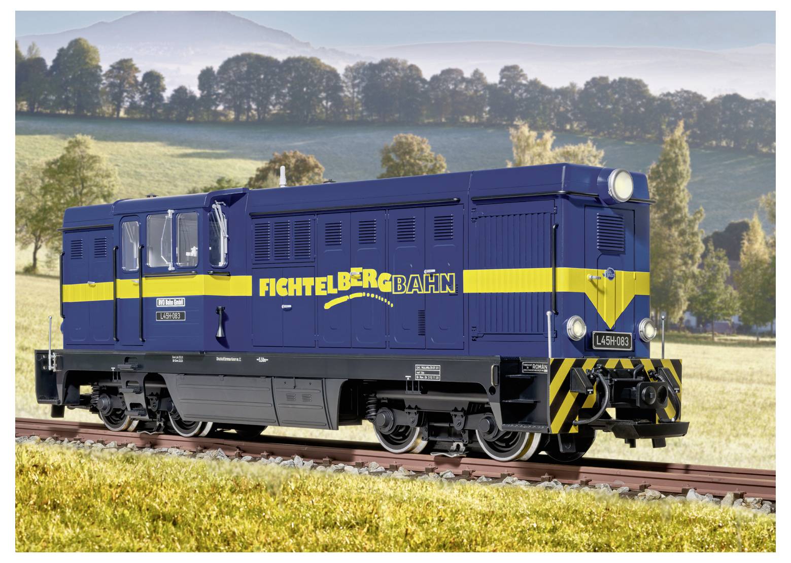 Blau-gelbe Fichtelbergbahn-Diesellokomotive auf Eisenbahngleisen in einer grasbewachsenen ländlichen Landschaft mit Hügeln und Bäumen im Hintergrund.