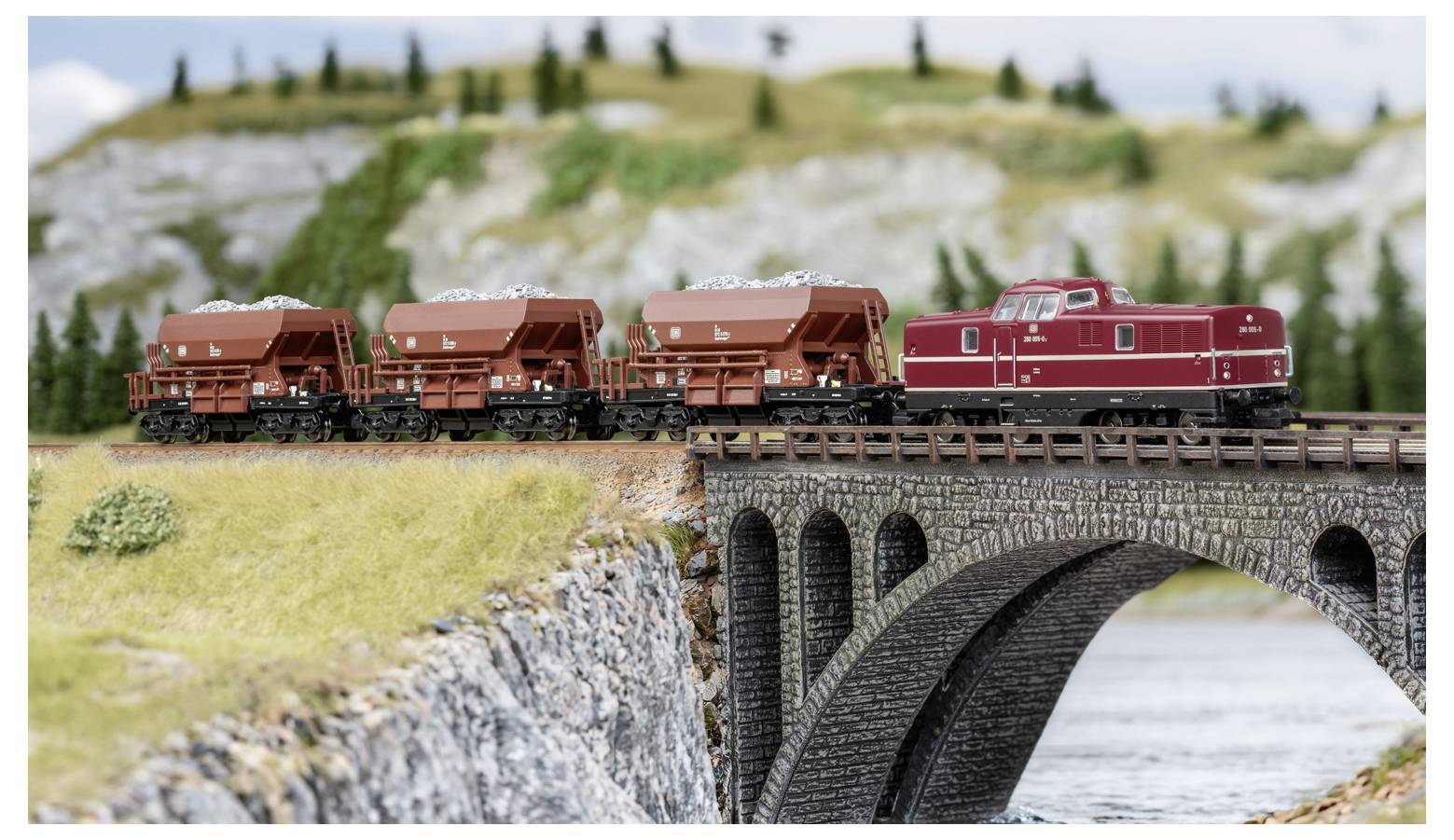 Modelleisenbahn mit rote Lokomotive und Güterwagen, die eine Steinbogenbrücke über Wasser in einer Miniaturlandschaft überquert.