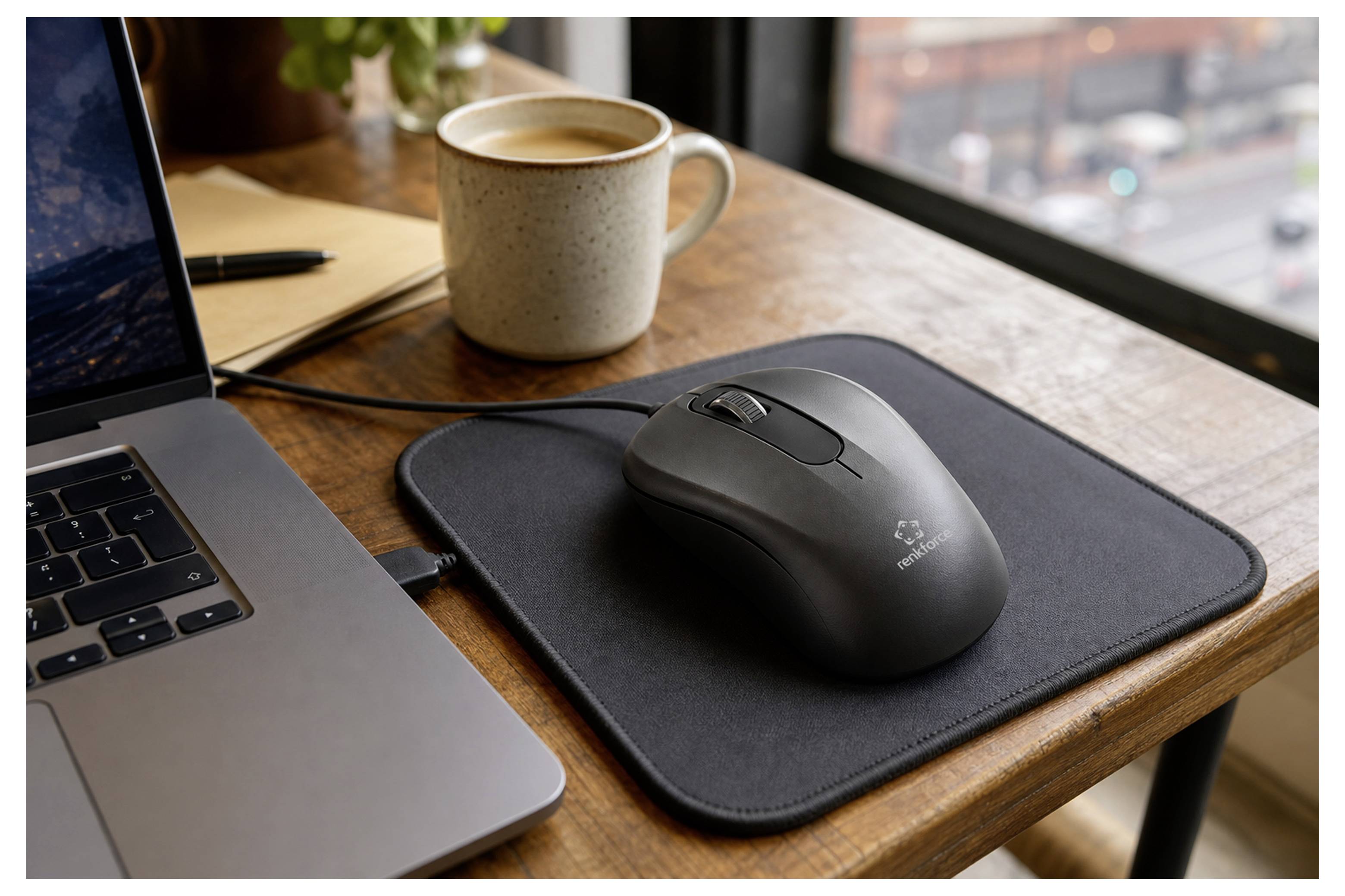 Kabellose Maus auf einem schwarzen Mousepad neben einem Laptop und einer Kaffeetasse auf einem Holzschreibtisch in der Nähe eines Fensters.