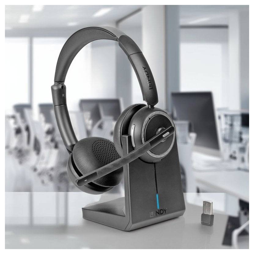 Schwarzes On-Ear-Headset mit Antennen-Mikrofon auf einer Ladestation, gezeigt auf einem Schreibtisch in einer unscharfen Büroumgebung mit einem USB-Dongle daneben.
