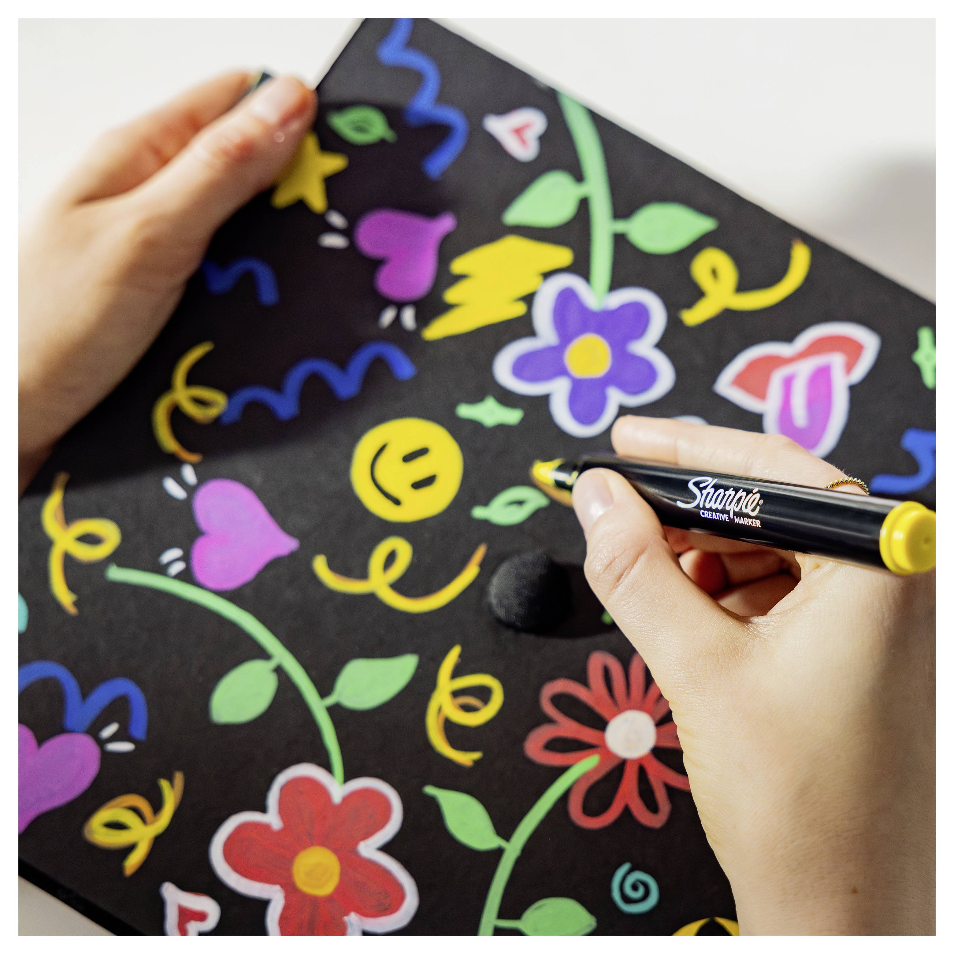 Hände zeichnen bunte Blumen, Herzen und lachende Gesichter auf schwarzes Papier mit einem gelb gespitzten Marker.