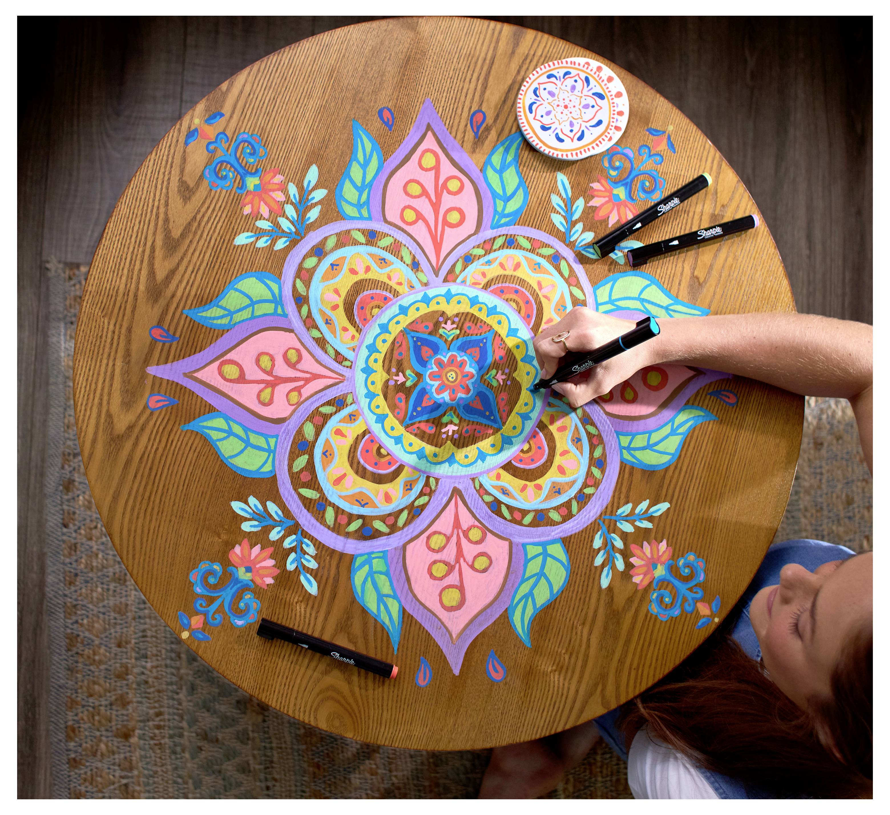 Person, die ein buntes Mandala mit Markern auf einem runden Holztisch zeichnet, von oben betrachtet.