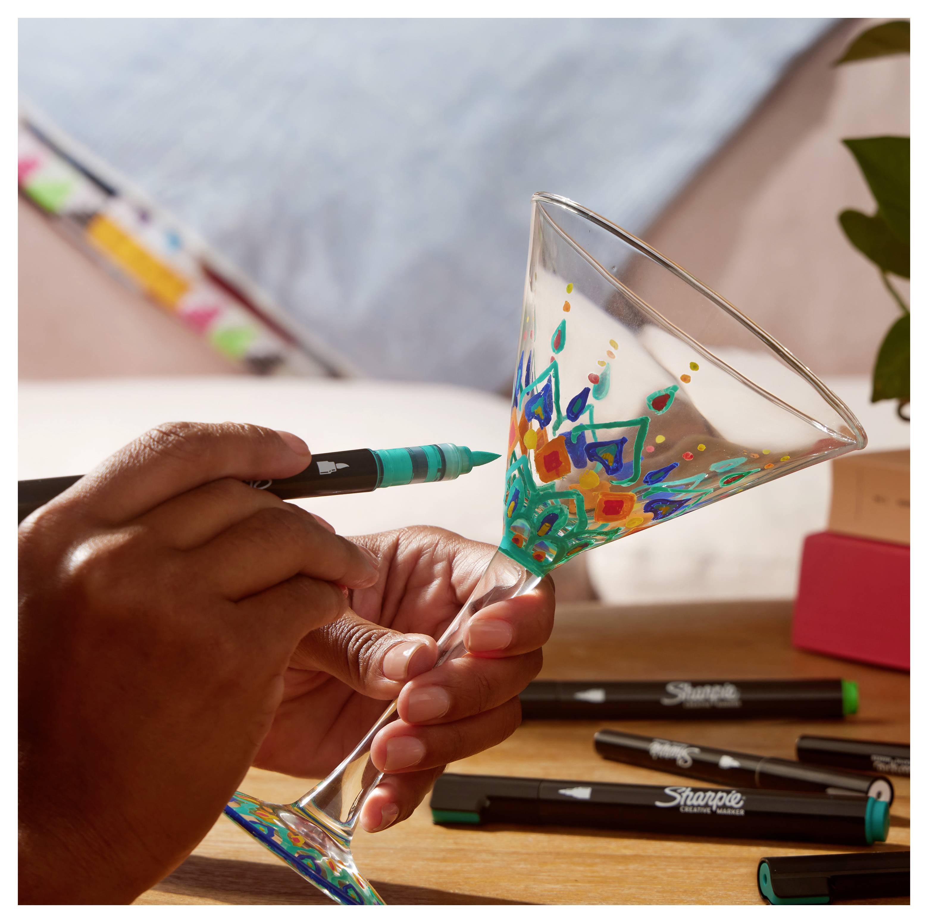 Eine Person dekoriert ein Martiniglas mit einem Blaugrünstift und zeichnet ein farbiges Blumenmuster auf das gestielte Glas.