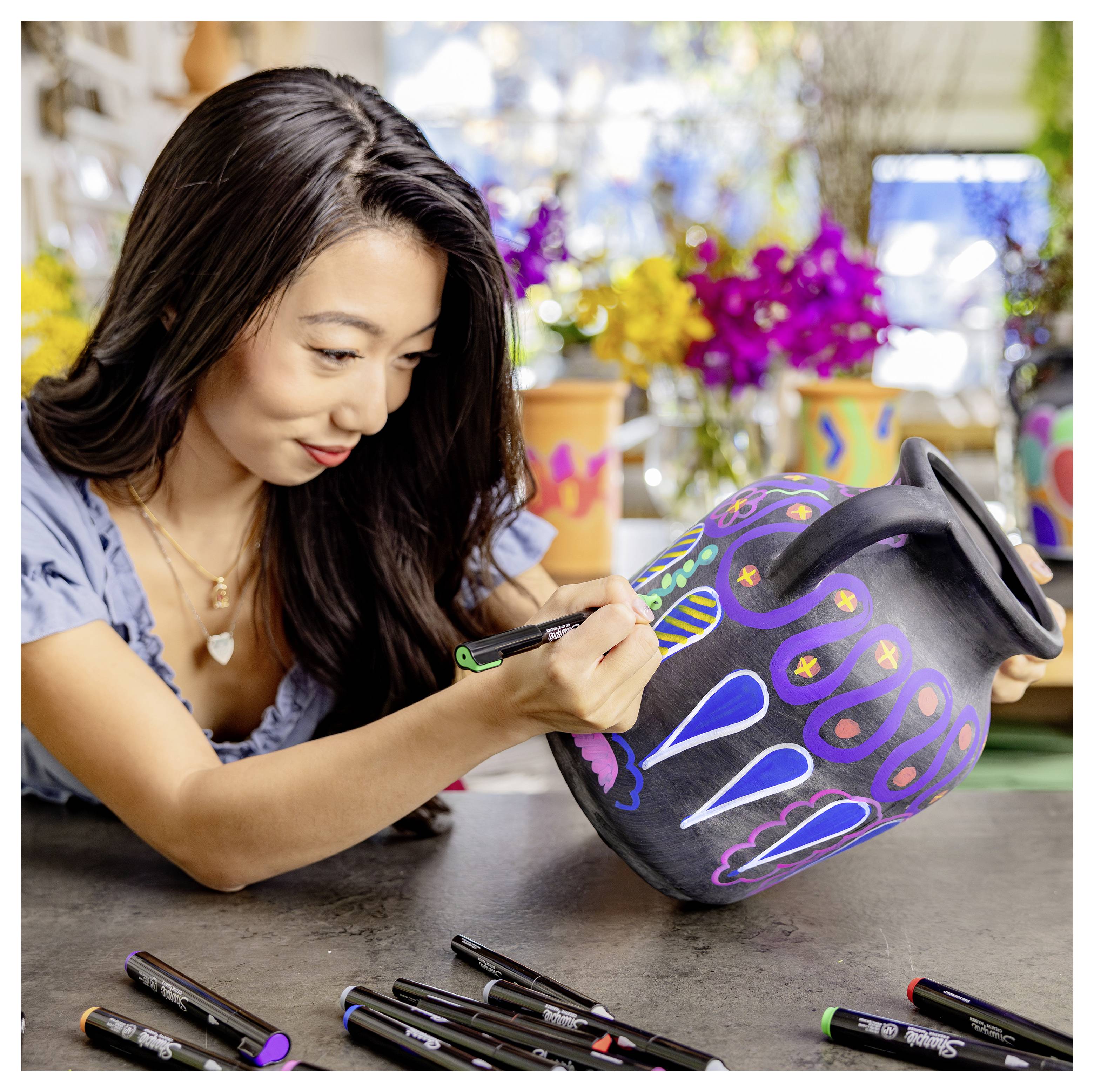 Frau, die eine schwarze Keramikvase mit bunten Blüten- und geometrischen Mustern mit Markern an einem Basteltisch verziert.