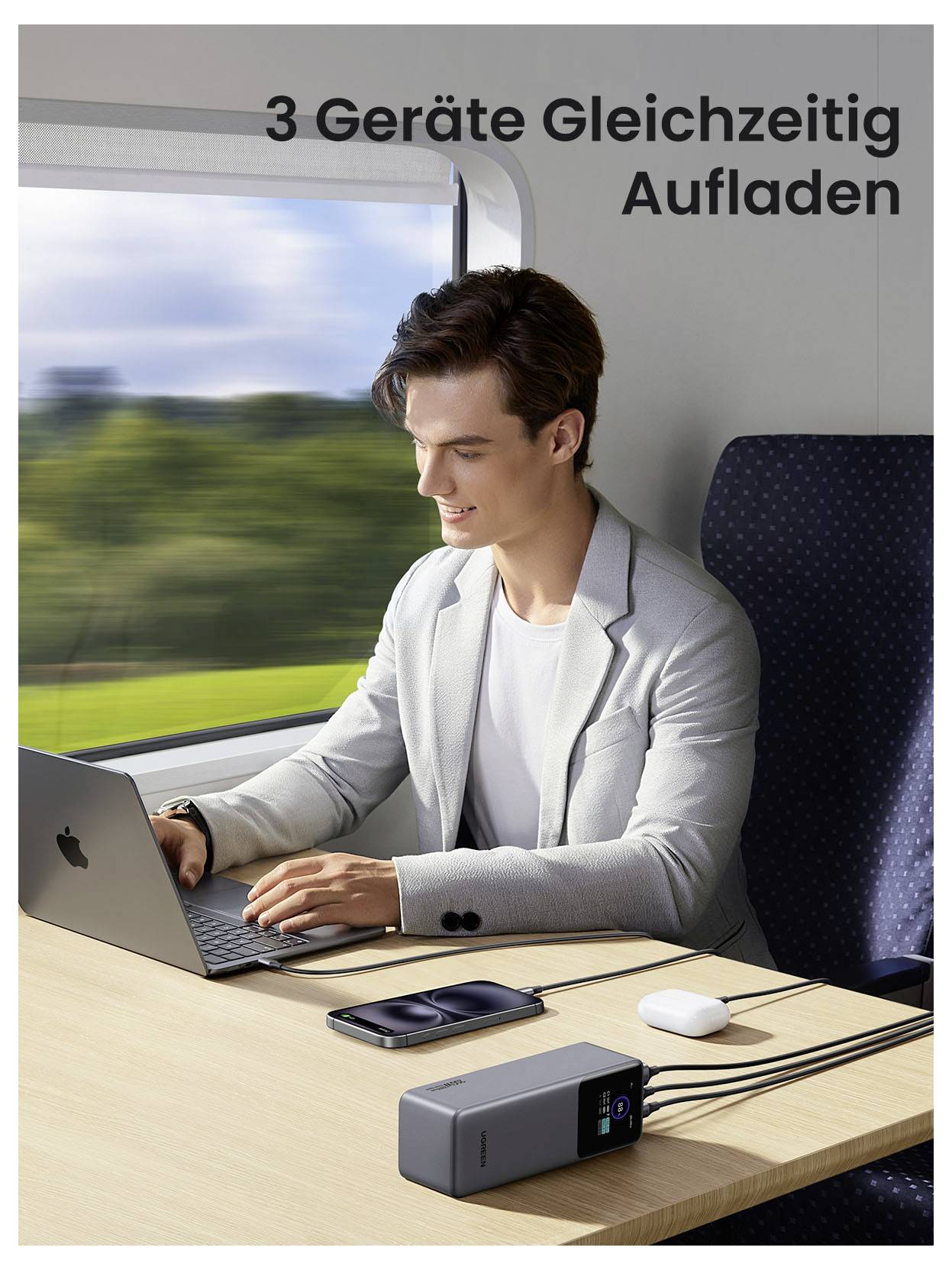 Ein Mann benutzt einen Laptop in einem Zug und lädt dabei gleichzeitig drei Geräte auf; deutscher Text lautet „3 Geräte Gleichzeitig Aufladen