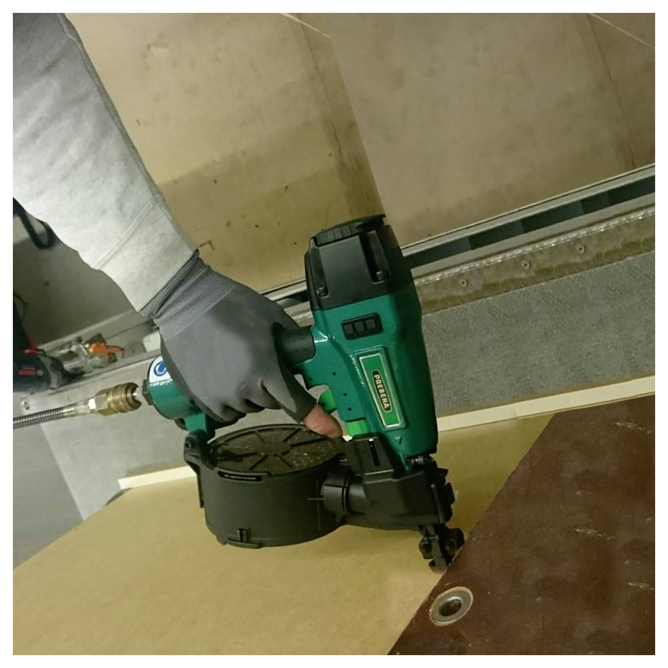 Behandschuhte Hand, die eine grüne kabellose Nagelmaschine verwendet, um ein Brett auf einer Arbeitsfläche zu befestigen.