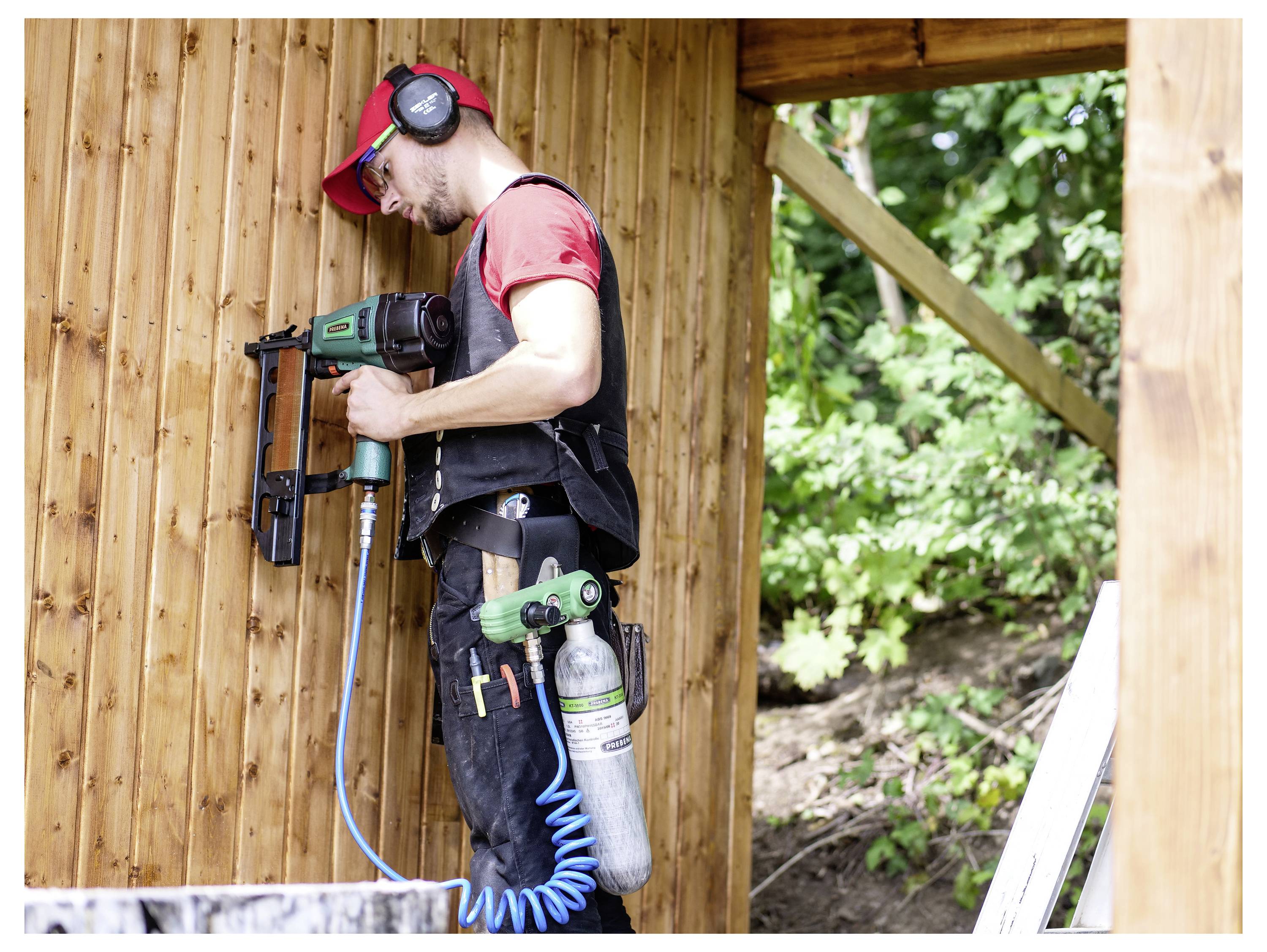Bauarbeiter benutzt eine pneumatische Nagelpistole, um Bretter an einer Holzwand zu befestigen, trägt Schutzausrüstung und hält einen Drucklufttank.