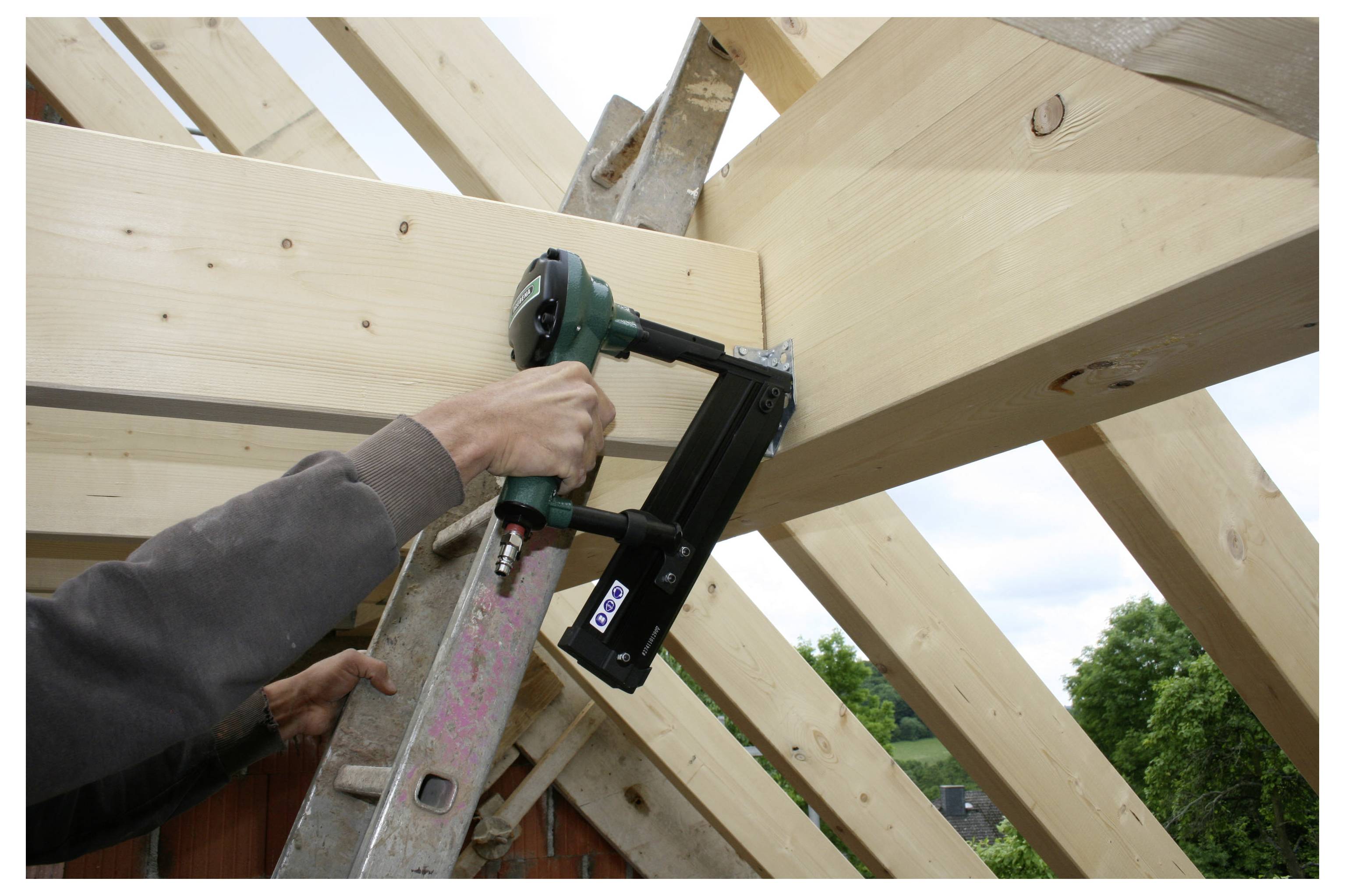 Arbeiter befestigt einen Holzbalken mit einem Nagelgewehr an der Dachkonstruktion, während er auf einer Leiter steht.