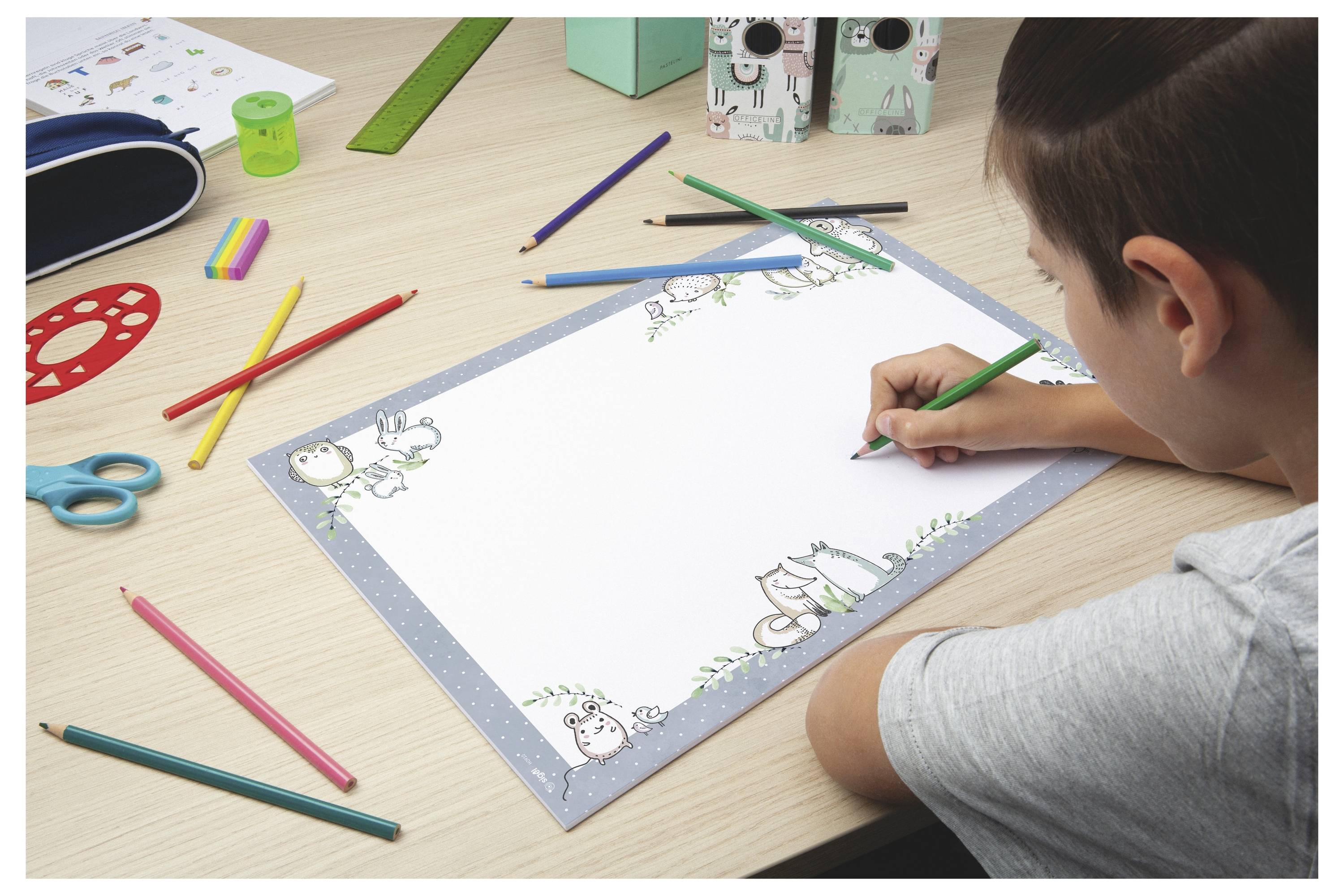 Kind malt ein leeres Arbeitsblatt mit Rahmen mit Buntstiften an einem Schreibtisch, umgeben von Schulmaterialien und Bastelbedarf.