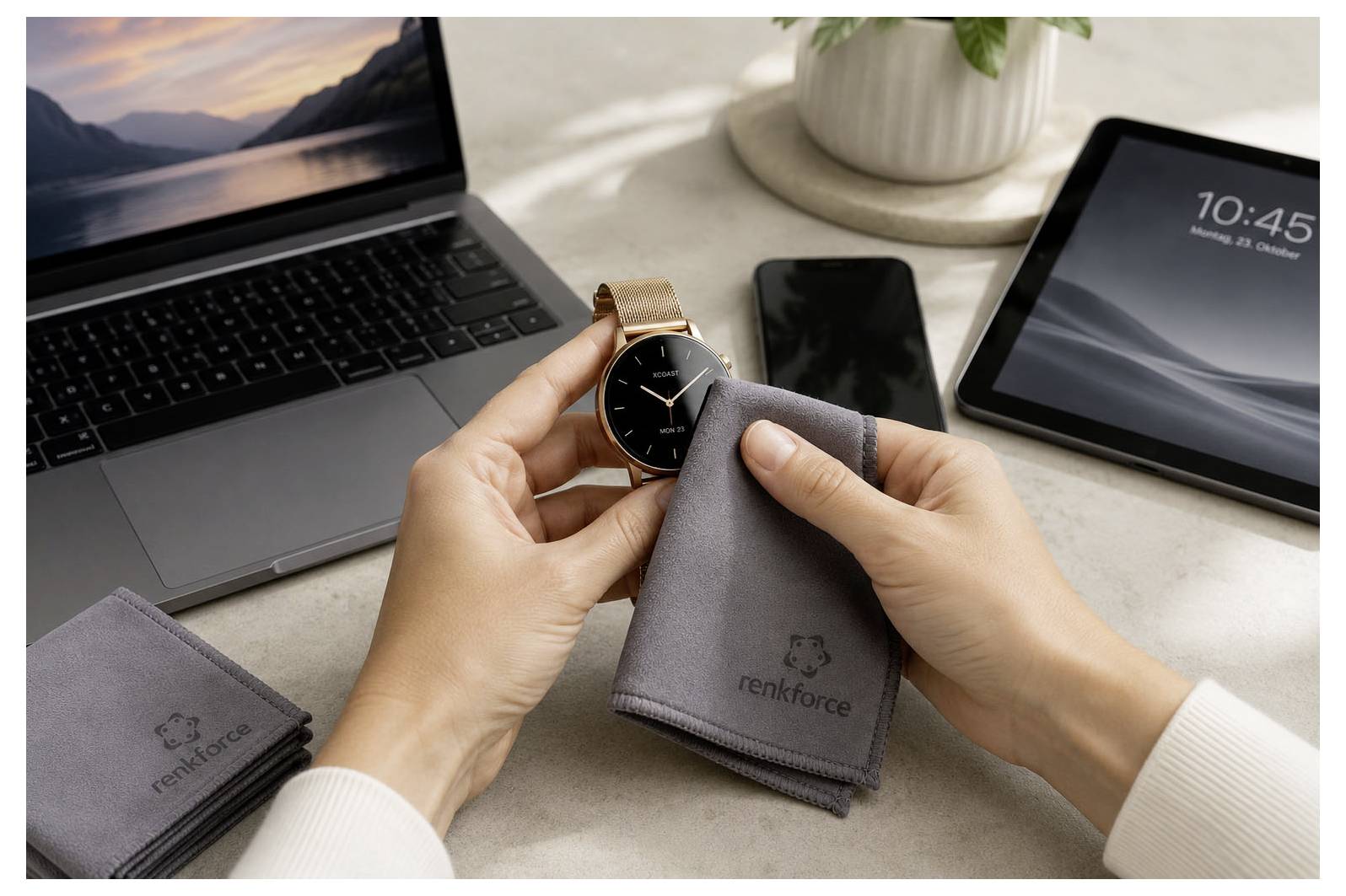 Hände, die eine Smartwatch und ein Reinigungstuch neben einem Laptop, Telefon und Tablet auf einem Schreibtisch halten, was auf die Reinigung von Bildschirmen oder Geräten hindeutet.
