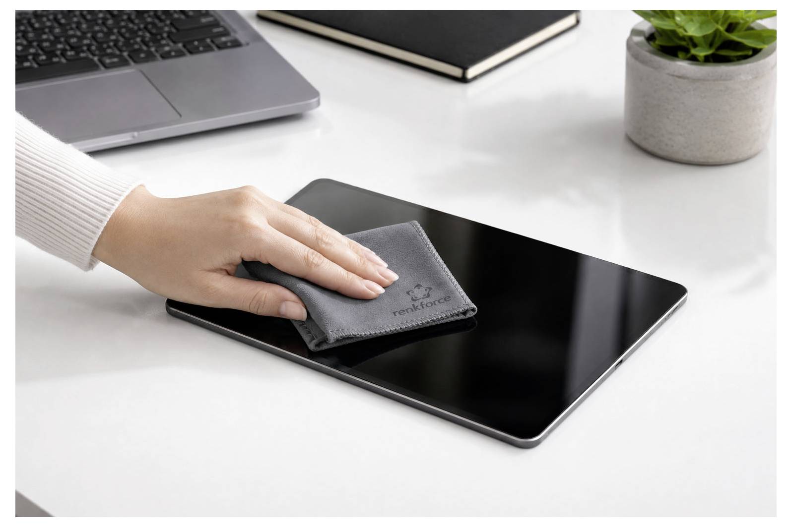 Eine Hand wischt einen schwarzen Tablet-Bildschirm mit einem grauen Tuch auf einem Schreibtisch neben einem Laptop und einer Pflanze ab.