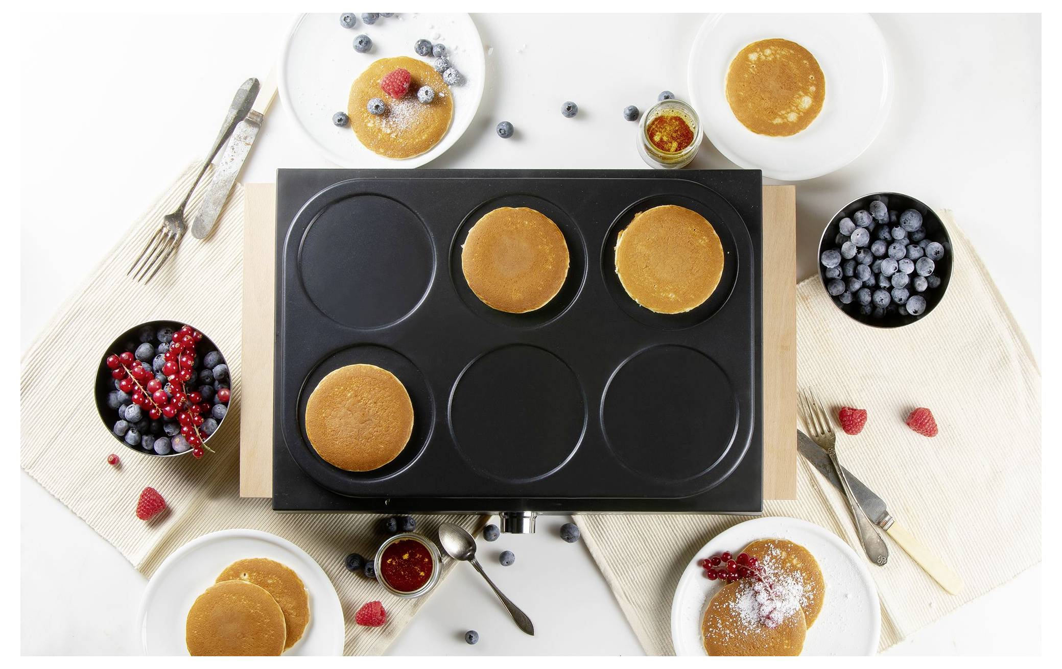 Draufsicht auf einen Pfannkuchenmacher mit vier kochenden Pfannkuchen, umgeben von Tellern mit Pfannkuchen, Beeren, Sirup und Utensilien auf einem weißen Tisch.