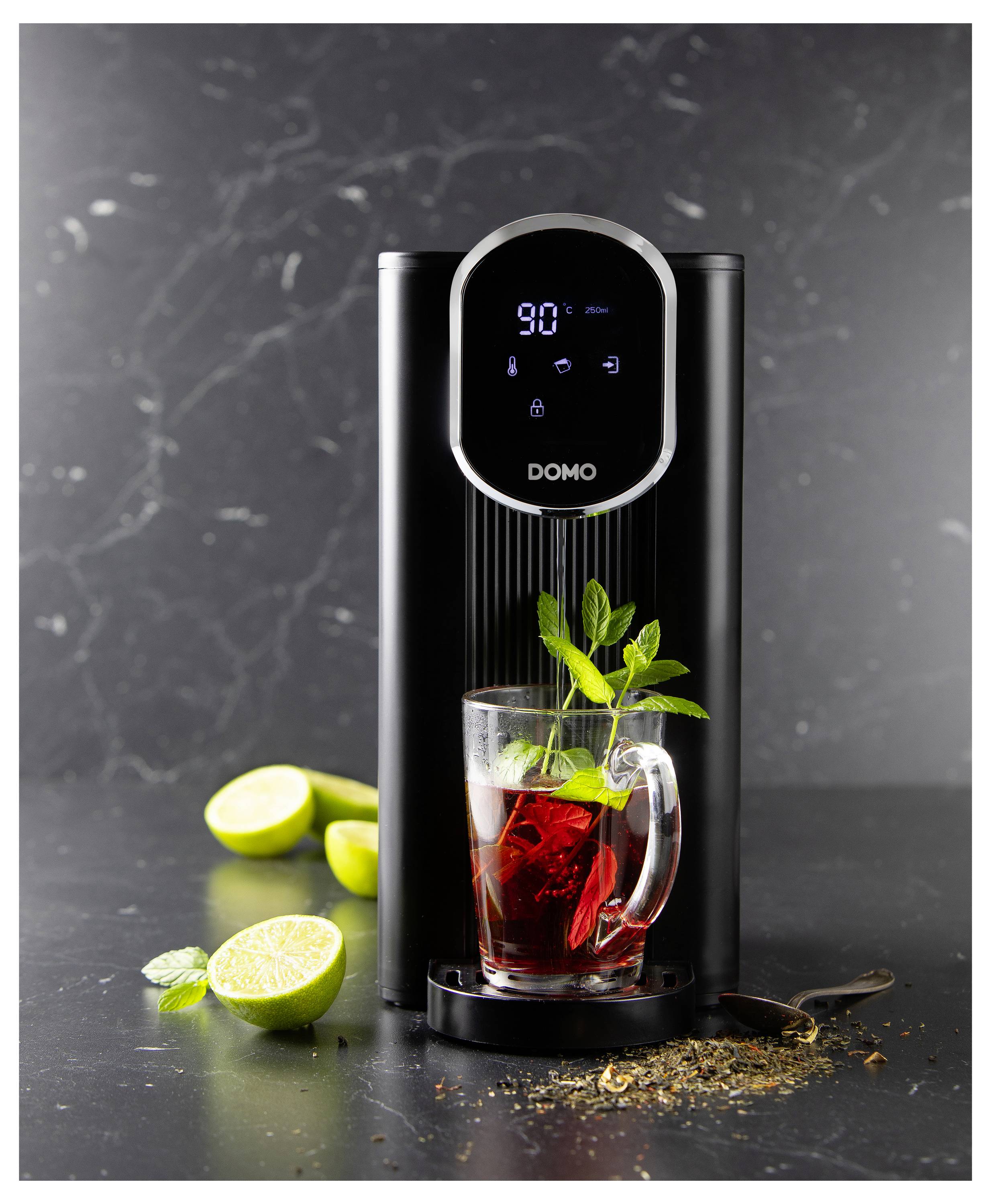 Schwarzer Domo-Heißwasserspender mit digitaler Anzeige; davor ein Glas mit rotem Tee, Minze und Limettenscheiben auf dunklem Hintergrund.