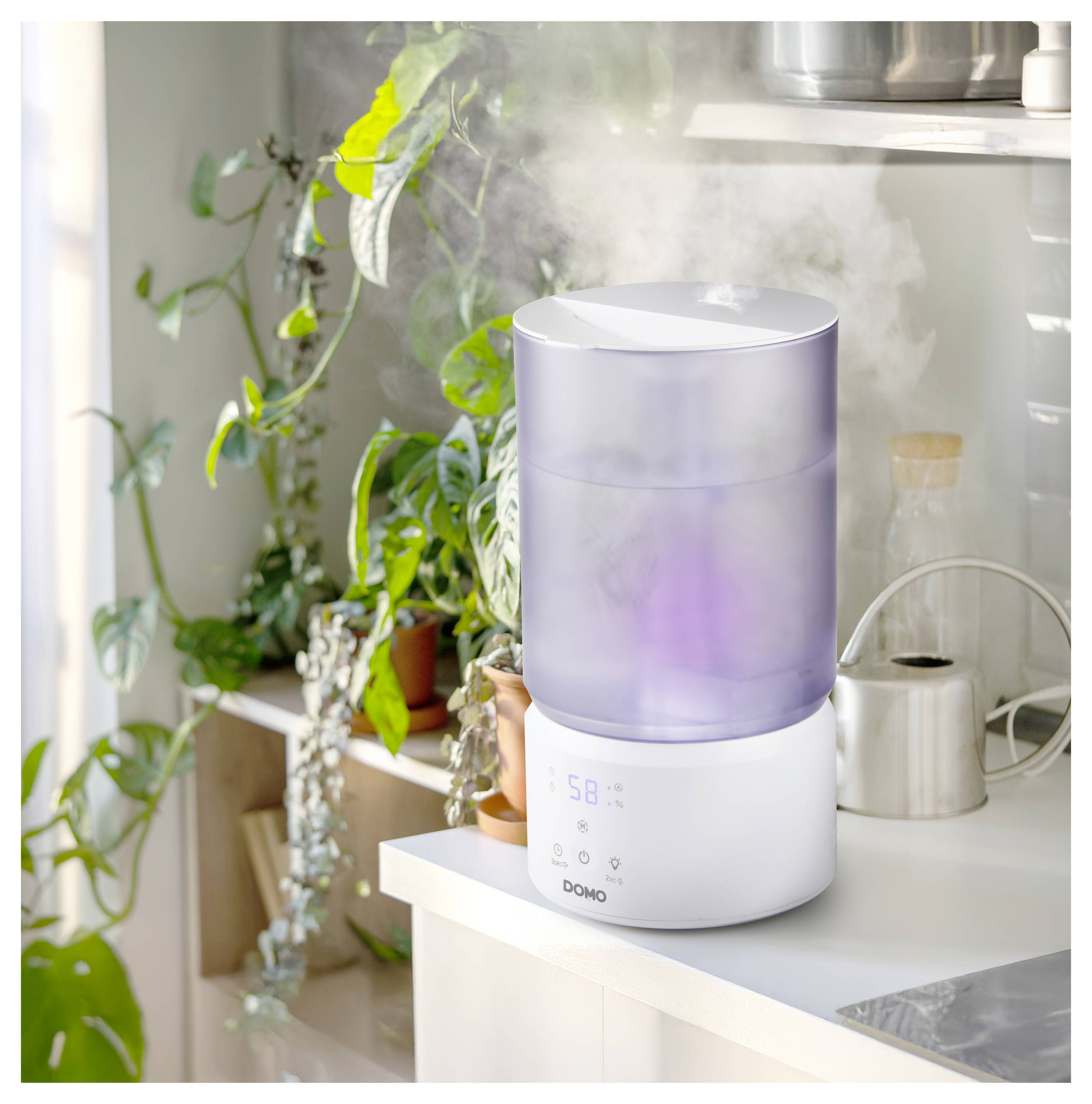 Ein weißer Ultraschall-Luftbefeuchter auf einer Küchentheke gibt Nebel neben Topfpflanzen und einer Gießkanne in einem hellen, sonnendurchfluteten Raum ab.