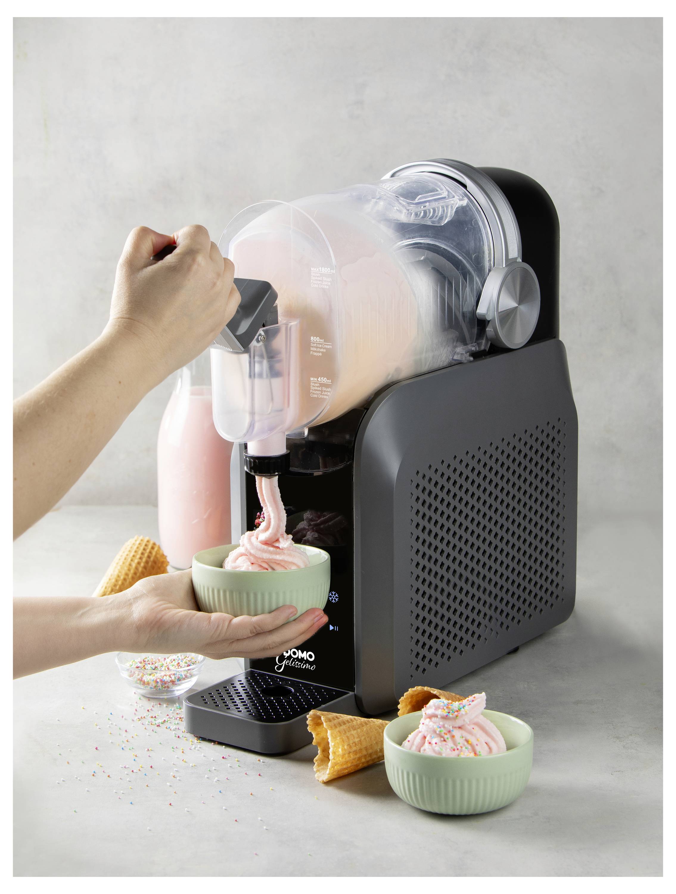 Eine Person verteilt rosafarbenes Softeis aus einer Eisthermaschine auf der Arbeitsfläche in eine Schale, wobei eine weitere gefüllte Schale in der Nähe steht und Waffeleistüten auf dem Tisch liegen.