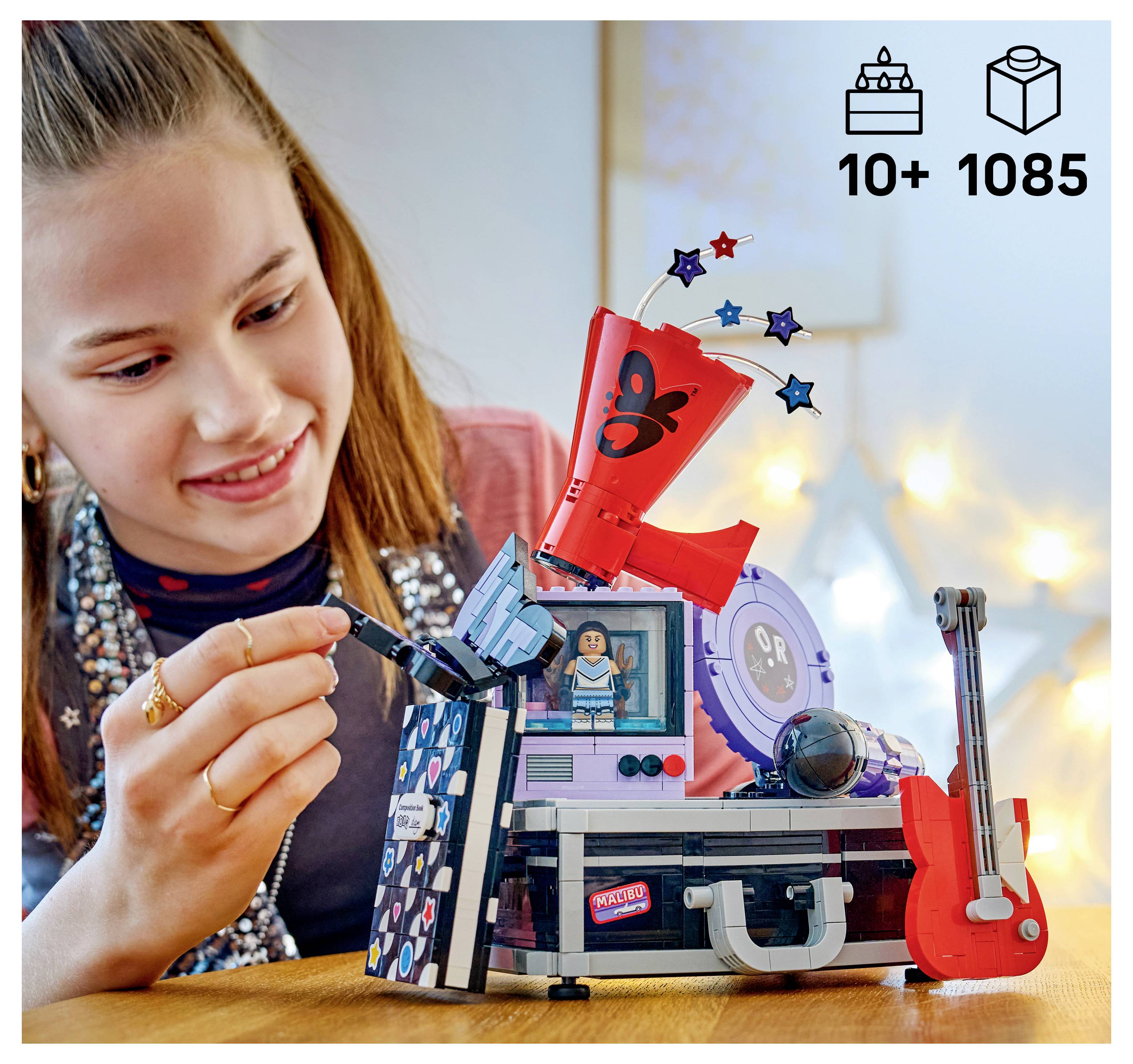Ein Mädchen baut ein LEGO-Musikspielset zusammen, das eine rote Gitarre, Bühnenscheinwerfer und eine Minifigur enthält; Symbole zeigen 10+ und 1085 Teile.
