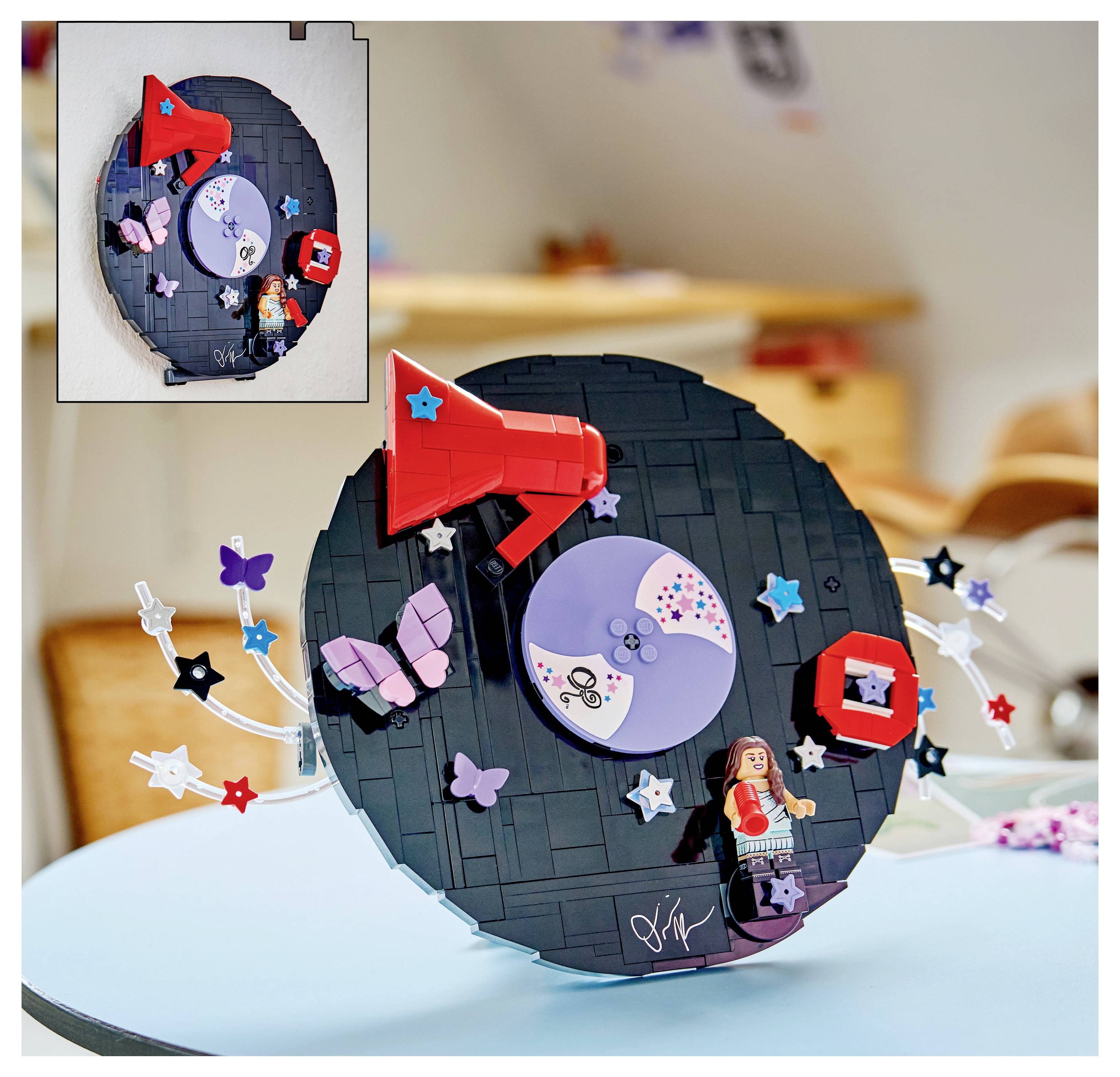 LEGO-artige zirkuläre Wandkunst mit einem roten Megafon, Schmetterlingen, Sternen und einer kleinen Minifigur auf einer hellblauen Tischplatte.