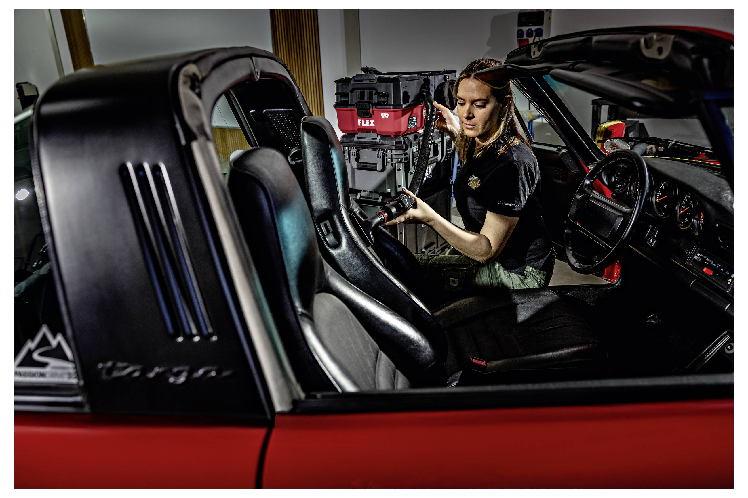 Ein Mechaniker arbeitet in einem roten Sportwagen und benutzt dabei Elektrowerkzeuge, während die Türen in einer Autowerkstatt offen sind.