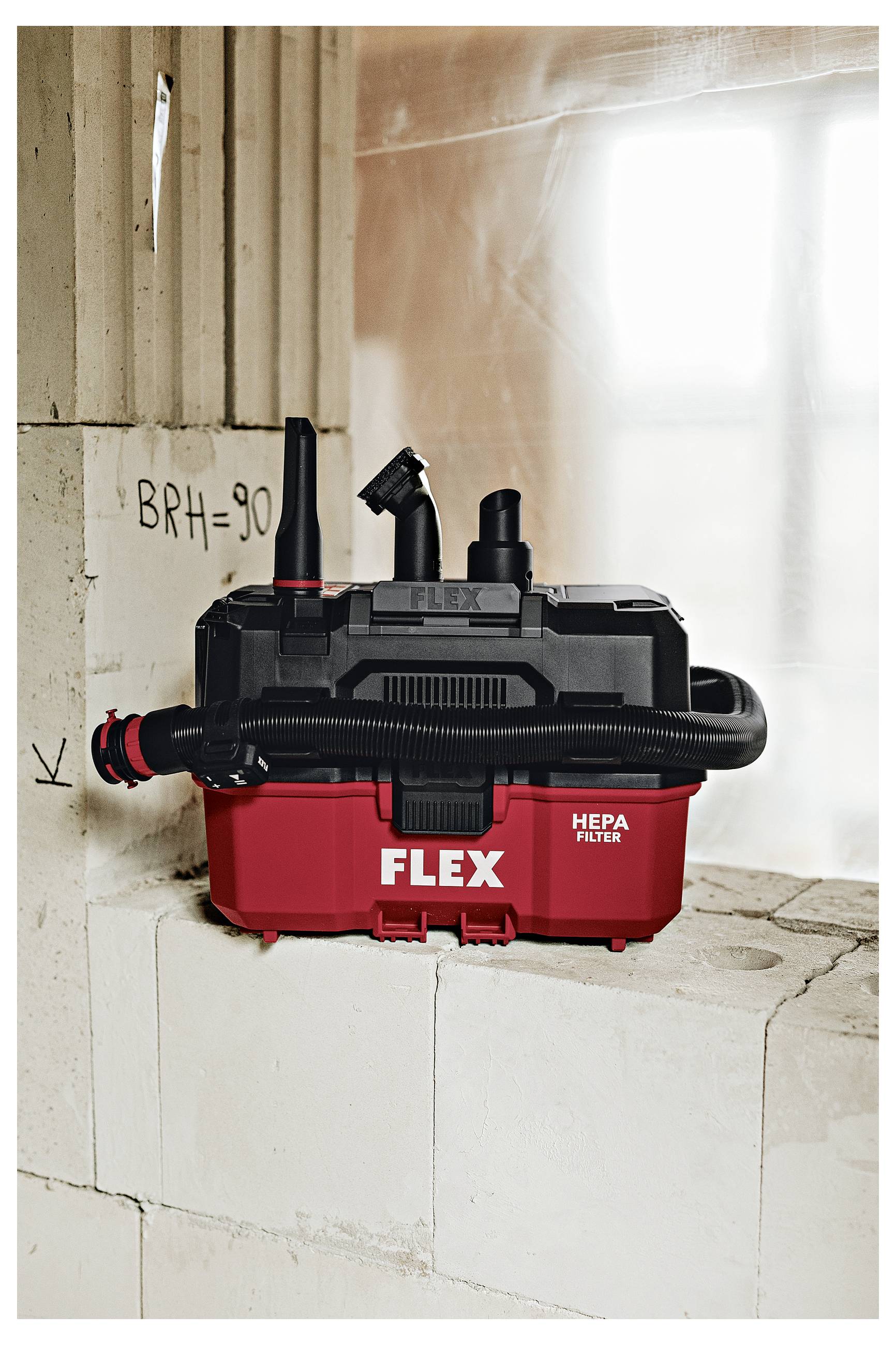 Roter FLEX HEPA-Staubsauger auf einer Baustelle, der auf gestapelten Betonsteinen neben unfertigen Wänden und einem hellen Fenster sitzt.