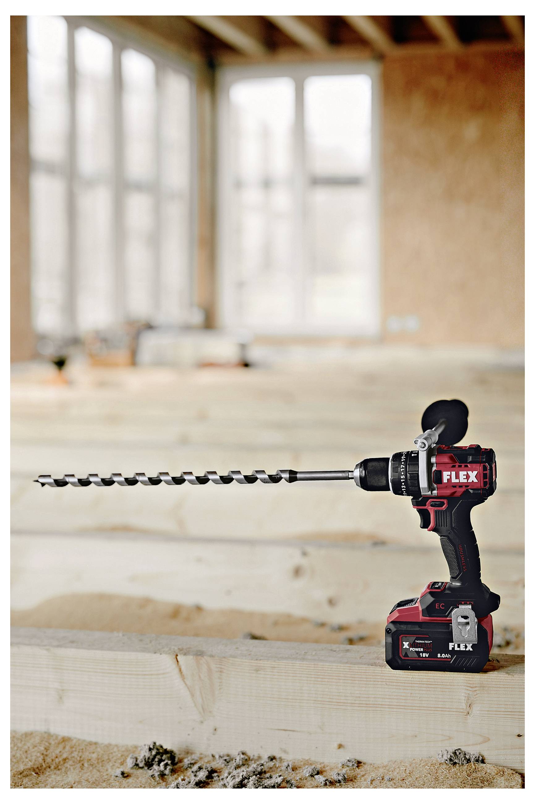 Kabelloses FLEX-Bohrgerät mit langem Schneckenbohreraufsatz, das auf einem Holzbalken in einem unfertigen Zimmer auf einer Baustelle ruht.