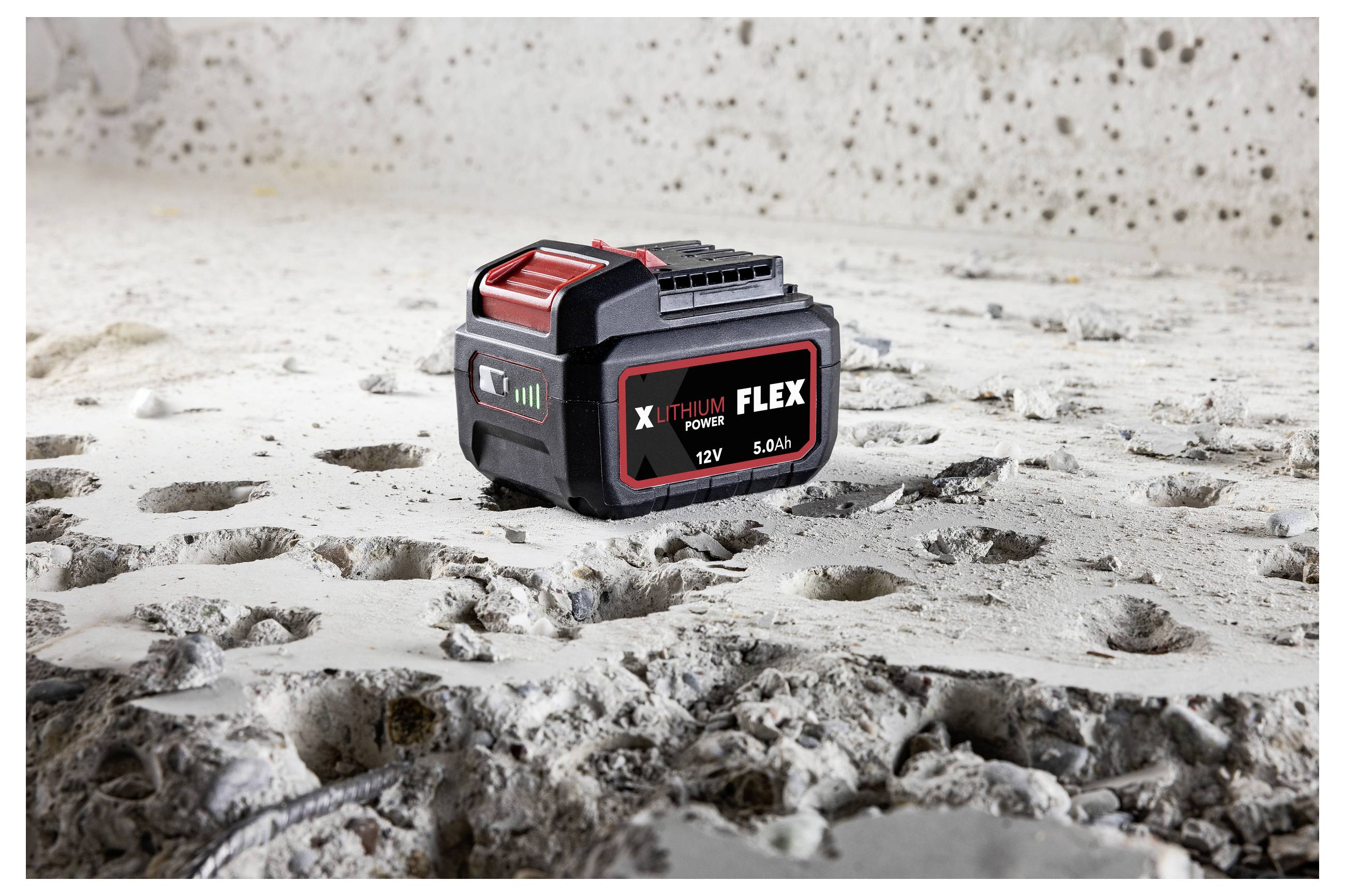 Schwarze und rote X-THIUM FLEX 12V 5,0Ah Elektrowerkzeug-Batterie, die auf einer staubigen, abgenutzten Betonoberfläche sitzt.