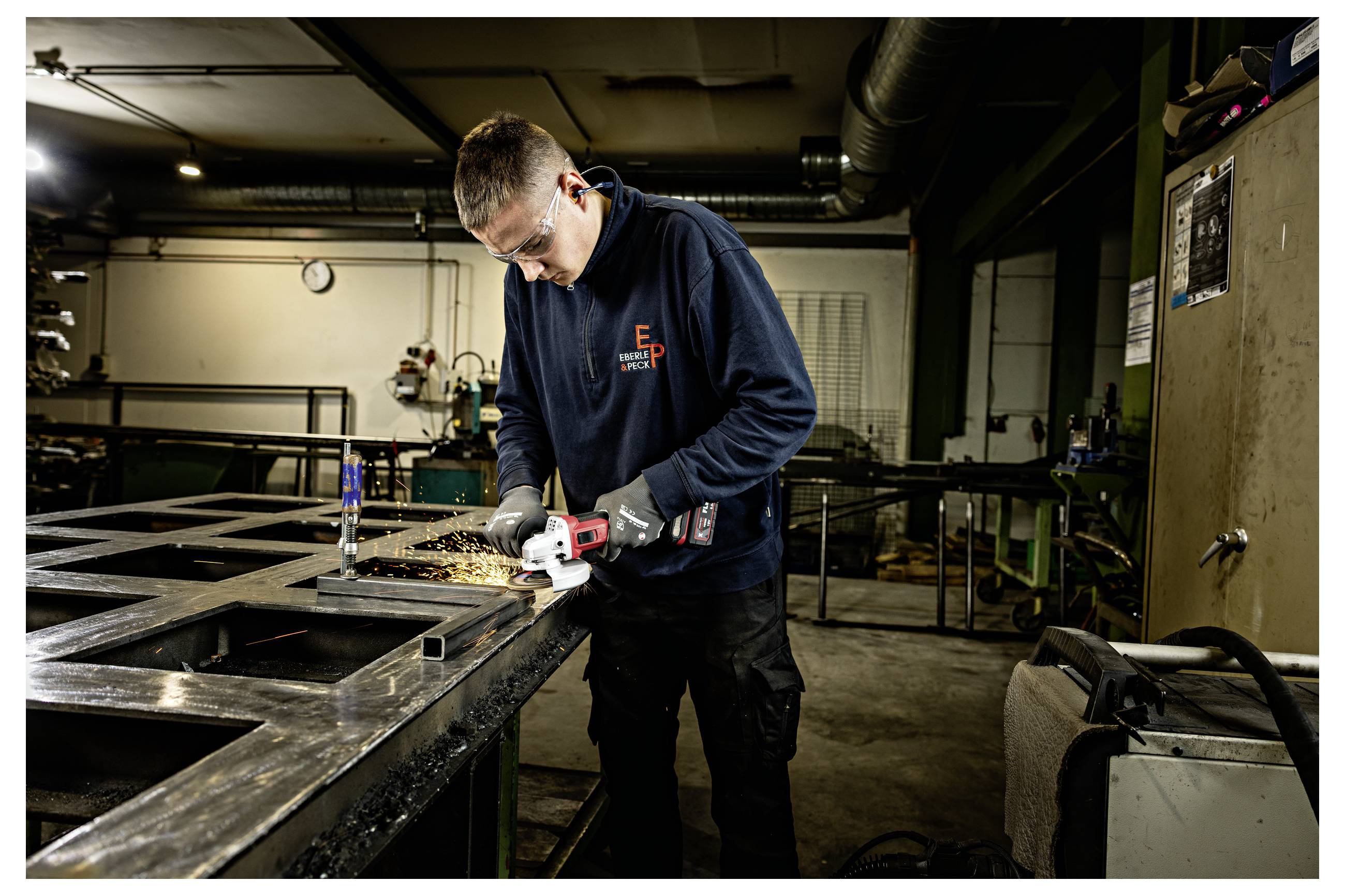 Ein Arbeiter in einer Werkstatt nutzt einen Winkelschleiffer an einem Metallrahmen, wobei Funken aus dem Schneidbereich fliegen.