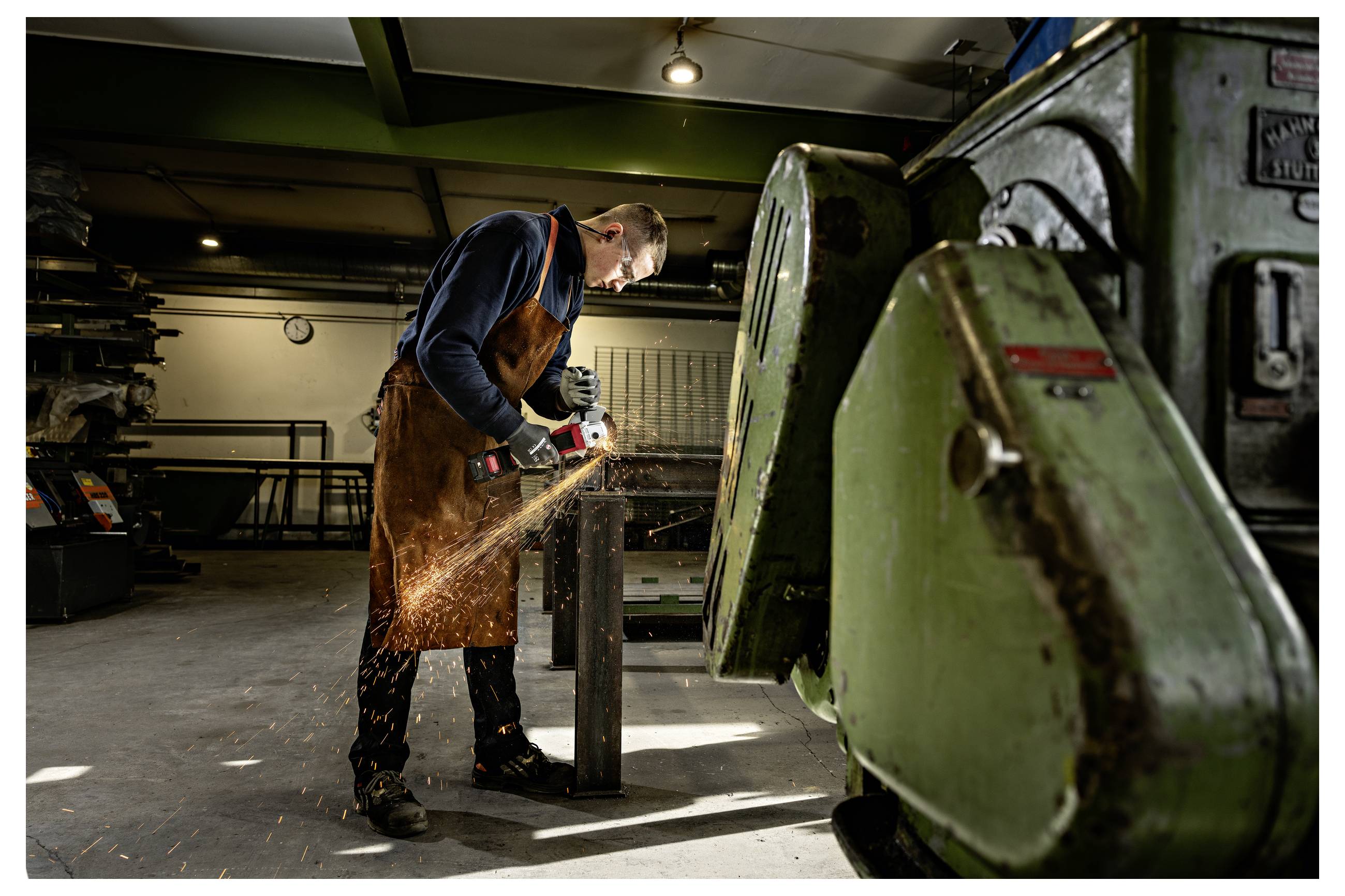 Metallarbeiter, der mit einem Winkelschleifer einen Metallpfosten in einer Werkstatt schneidet, mit fliegenden Funken und schweren Maschinen in der Nähe.