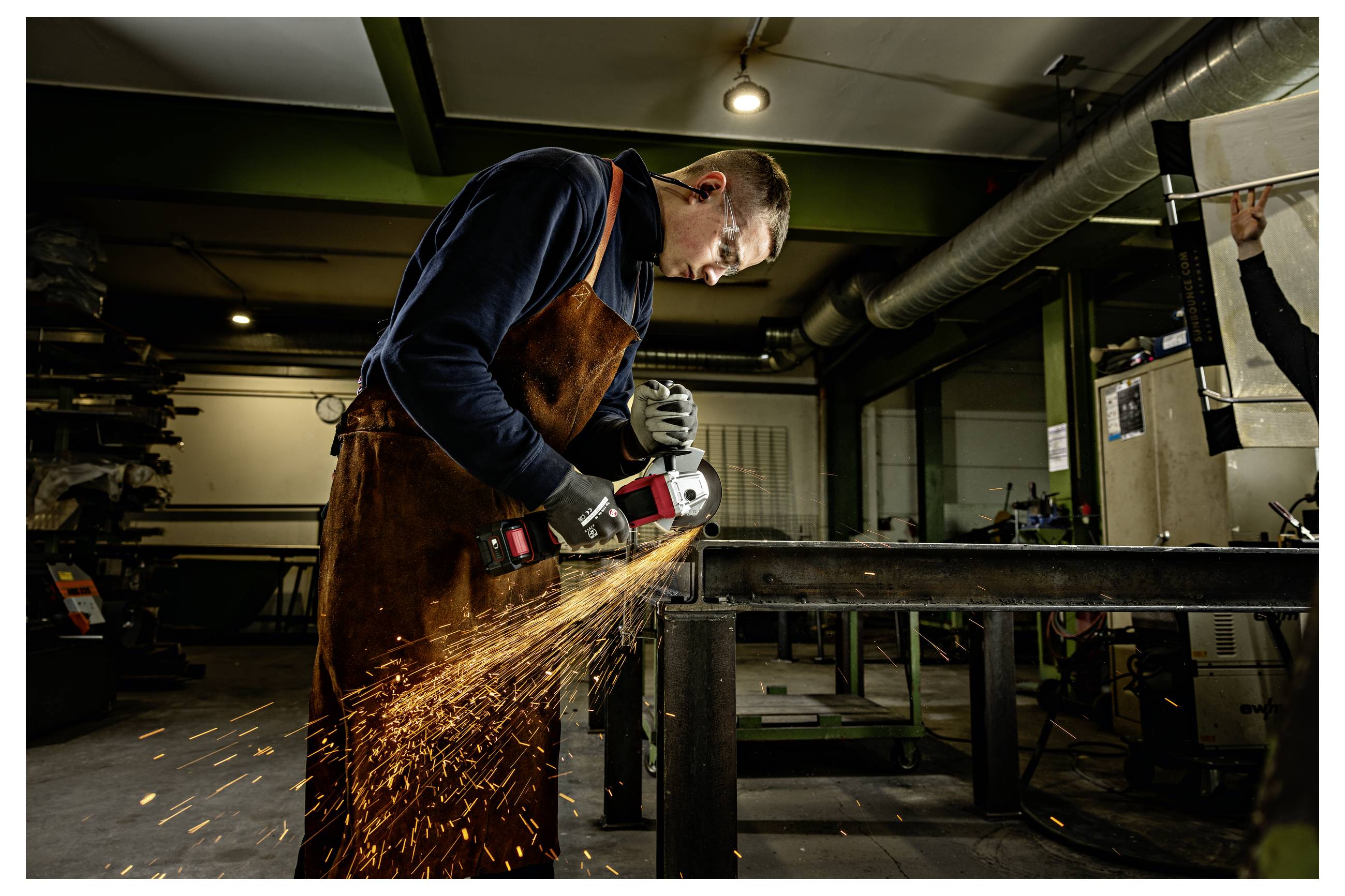 Ein Schweißer in einer Werkstatt verwendet einen Winkelschleifer an einem Metallrahmen und wirft dabei helle Funken auf.