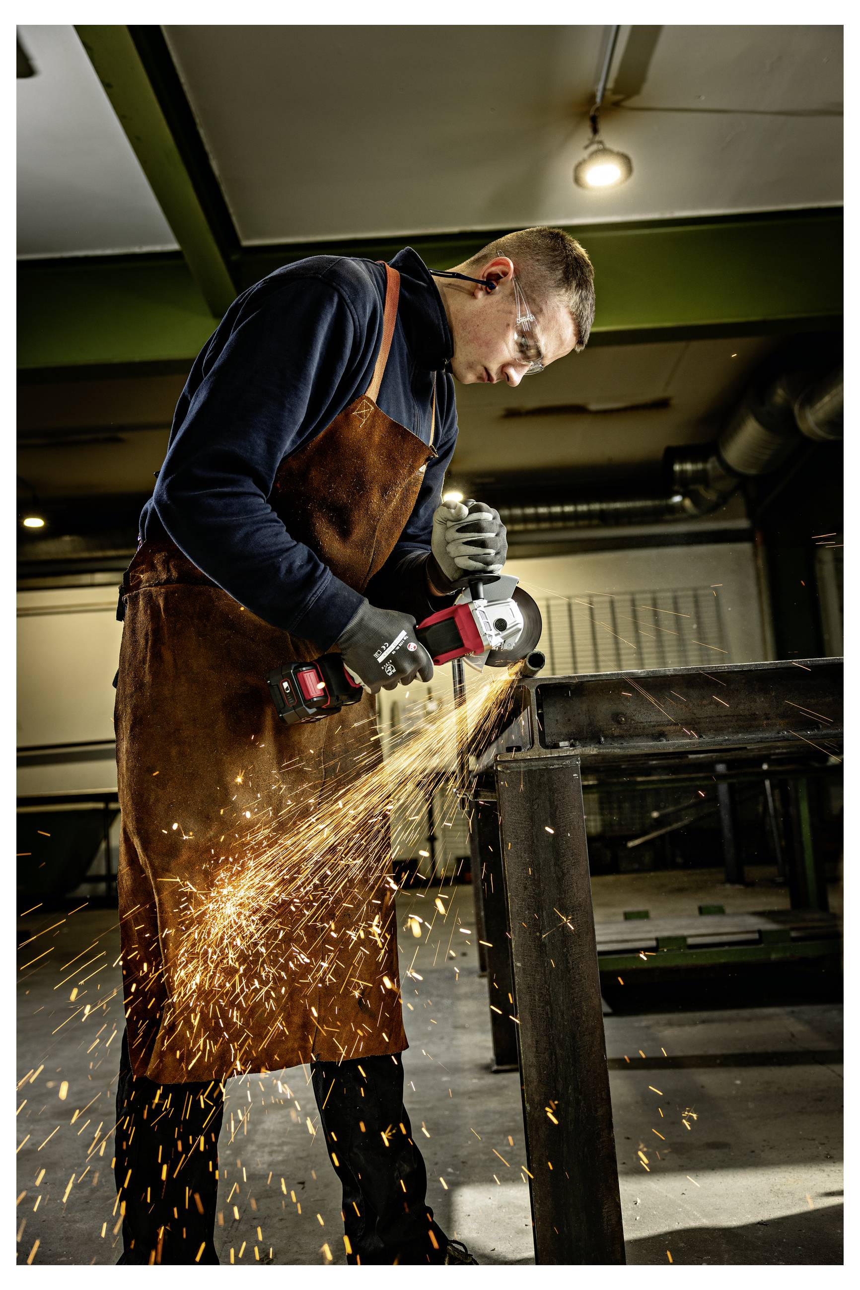 Mann in Schürze und Handschuhen, der einen Winkelschleiffer auf einem Metalltisch benutzt und dabei helle Funken in einer Werkstatt versendet.