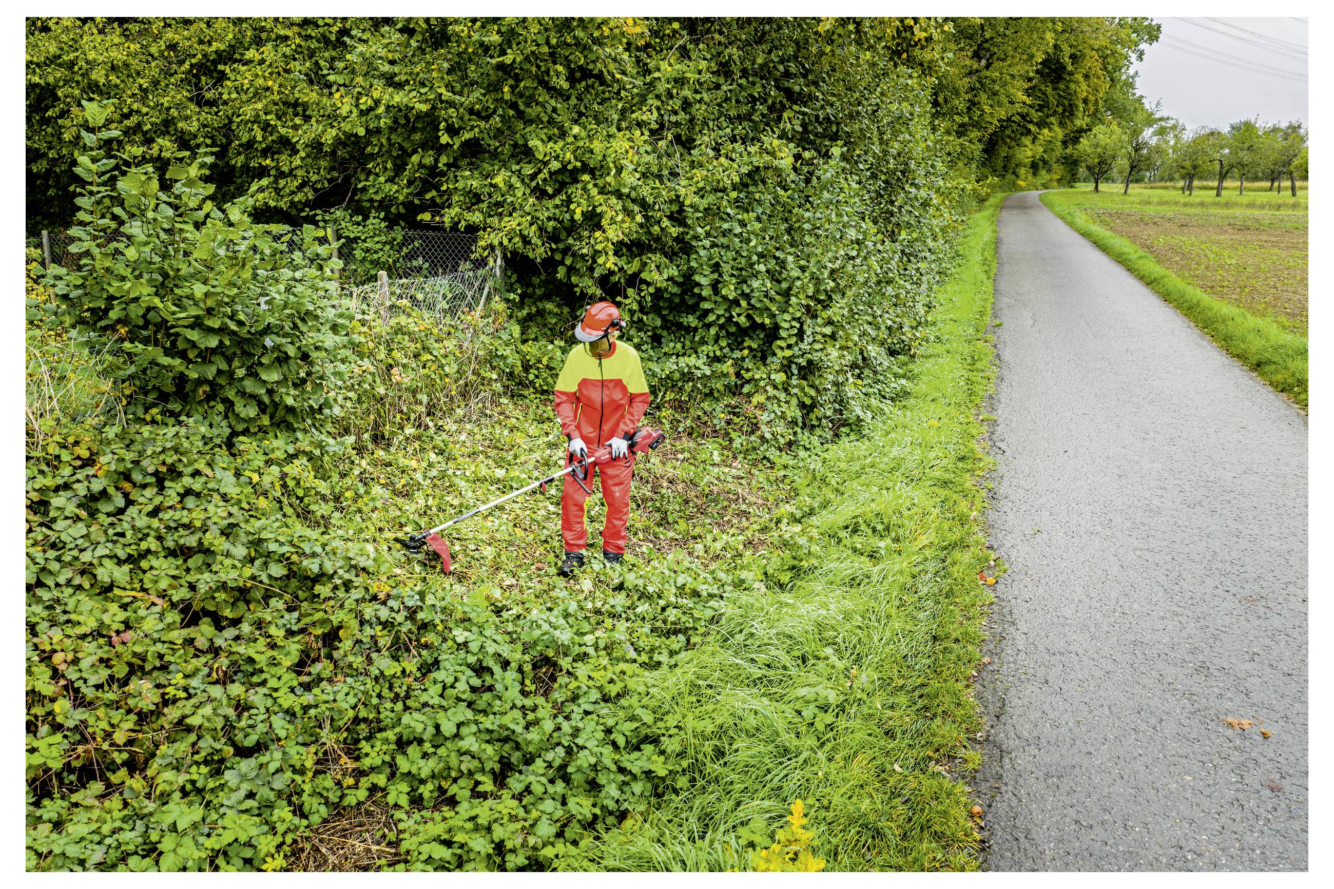 Ein Arbeiter in orangefarbener Schutzausrüstung verwendet einen Fadenmäher, um überwuchertes Straßenrandgestrüpp neben einer ländlichen asphaltierten Straße zu schneiden.
