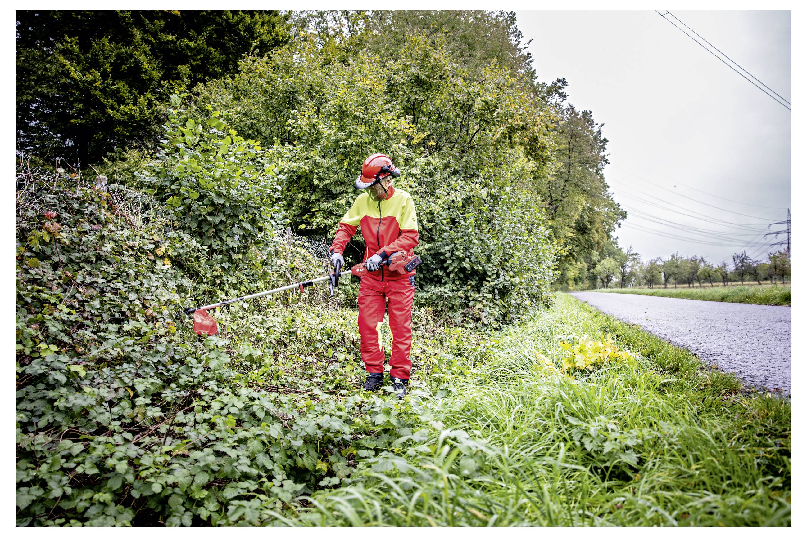 Arbeiter in roter Schutzausrüstung, der mit einem Fadentrimmer Straßenrandvegetation neben einer ländlichen Straße stutzt.