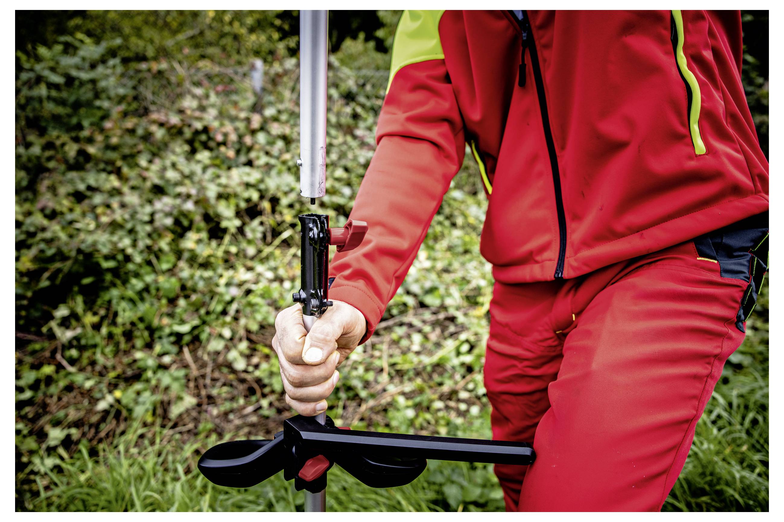 Nahaufnahme einer Person in roter Outdoor-Kleidung, die eine Stange oder ein Werkzeug mit einer schwarzen Klemme hält, im Freien in der Nähe von Gras und Sträuchern.