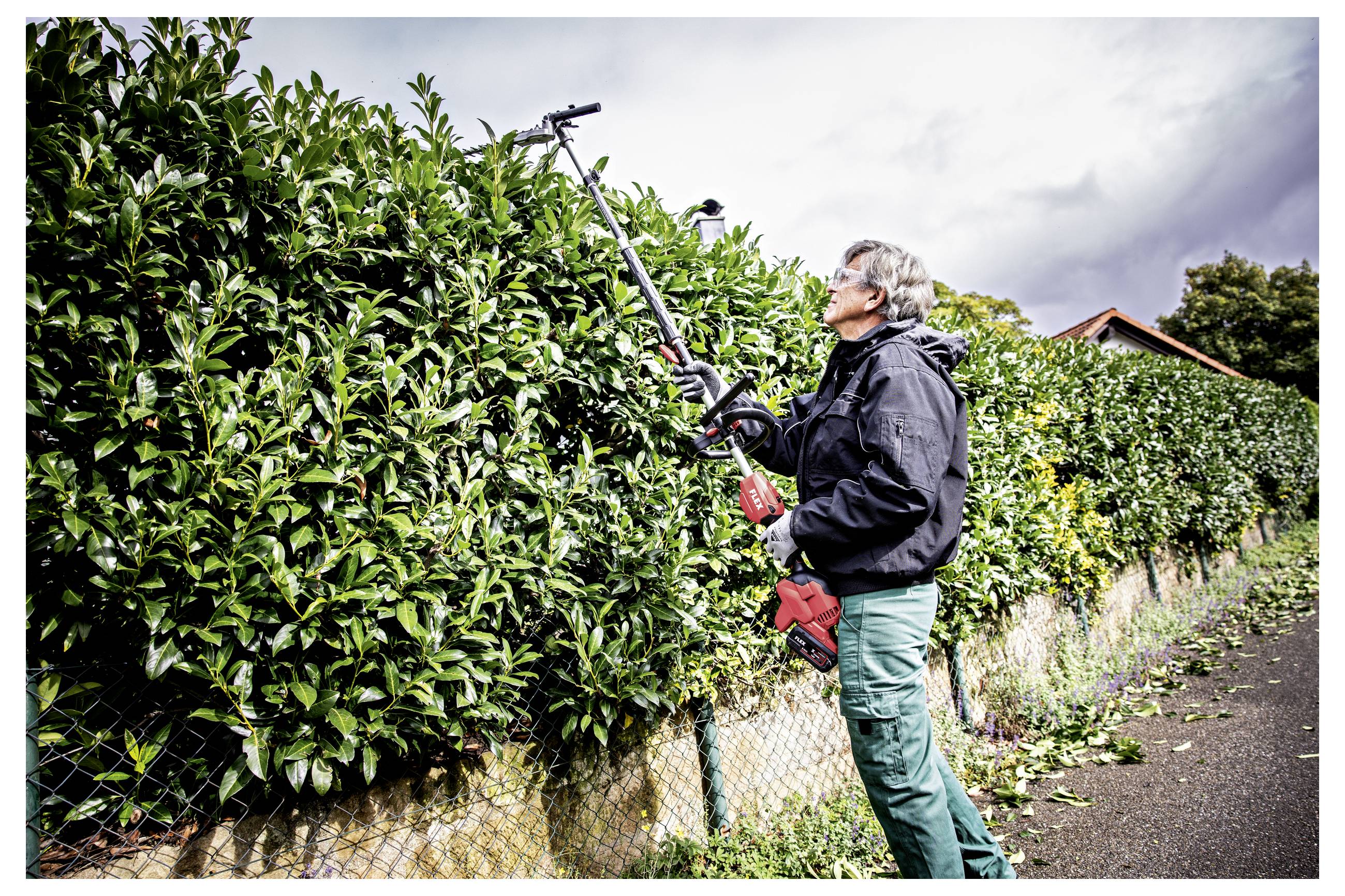 Person, die eine hohe Hecke mit einer Teleskopsäge entlang eines Straßenzaunzuschneidet.