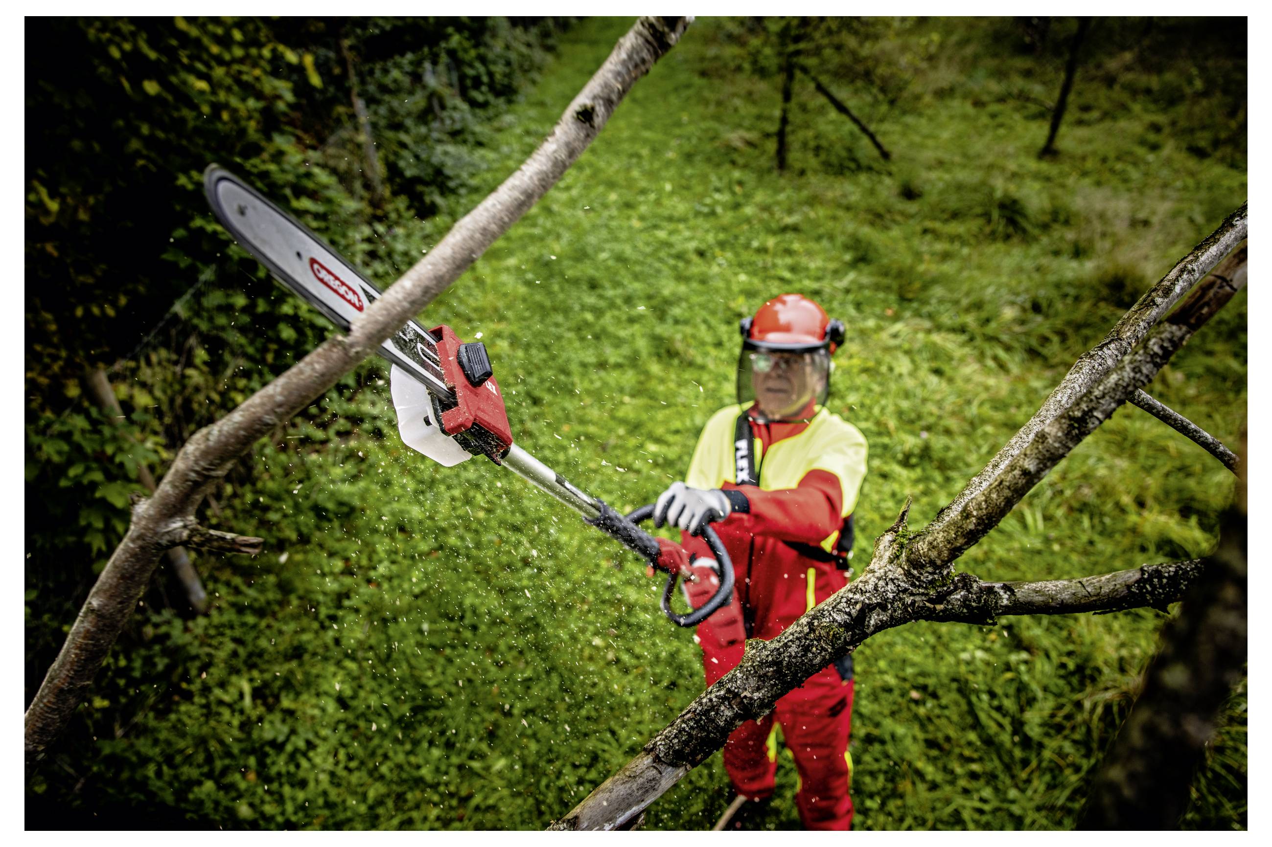 Arbeiter in roter Schutzausrüstung verwendet eine Kettensägen-Teleskopsäge zum Schneiden von Ästen von einem Baum an einem steilen Grashang.