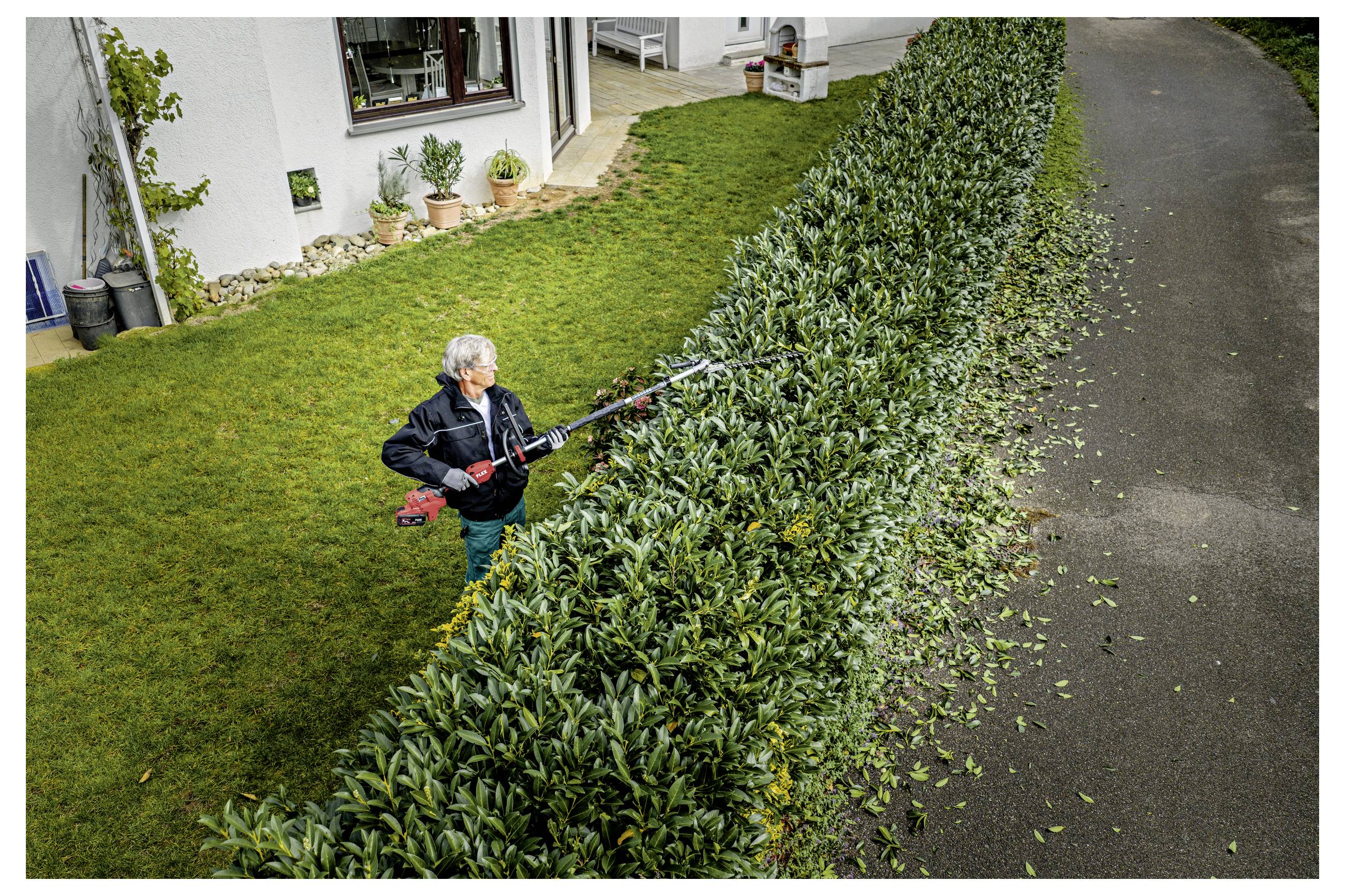 Person, die eine lange Hecke neben einem Haus mit einer Heckenschere schneidet, mit Schnittgut, das entlang der Straße verstreut ist.