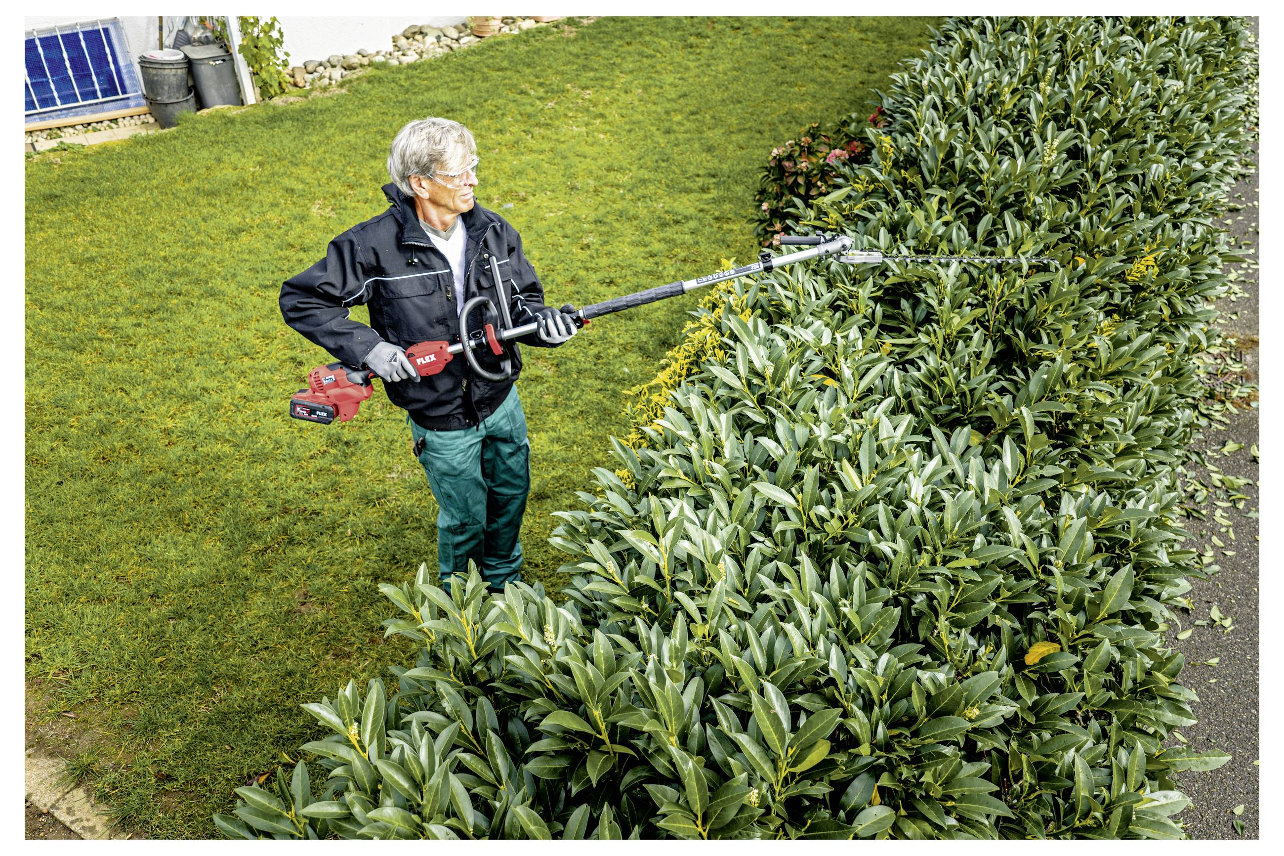 Älterer Mann, der eine Heckenschere verwendet, um eine große grüne Strauchbegrenzung in einem Garten zu schneiden.